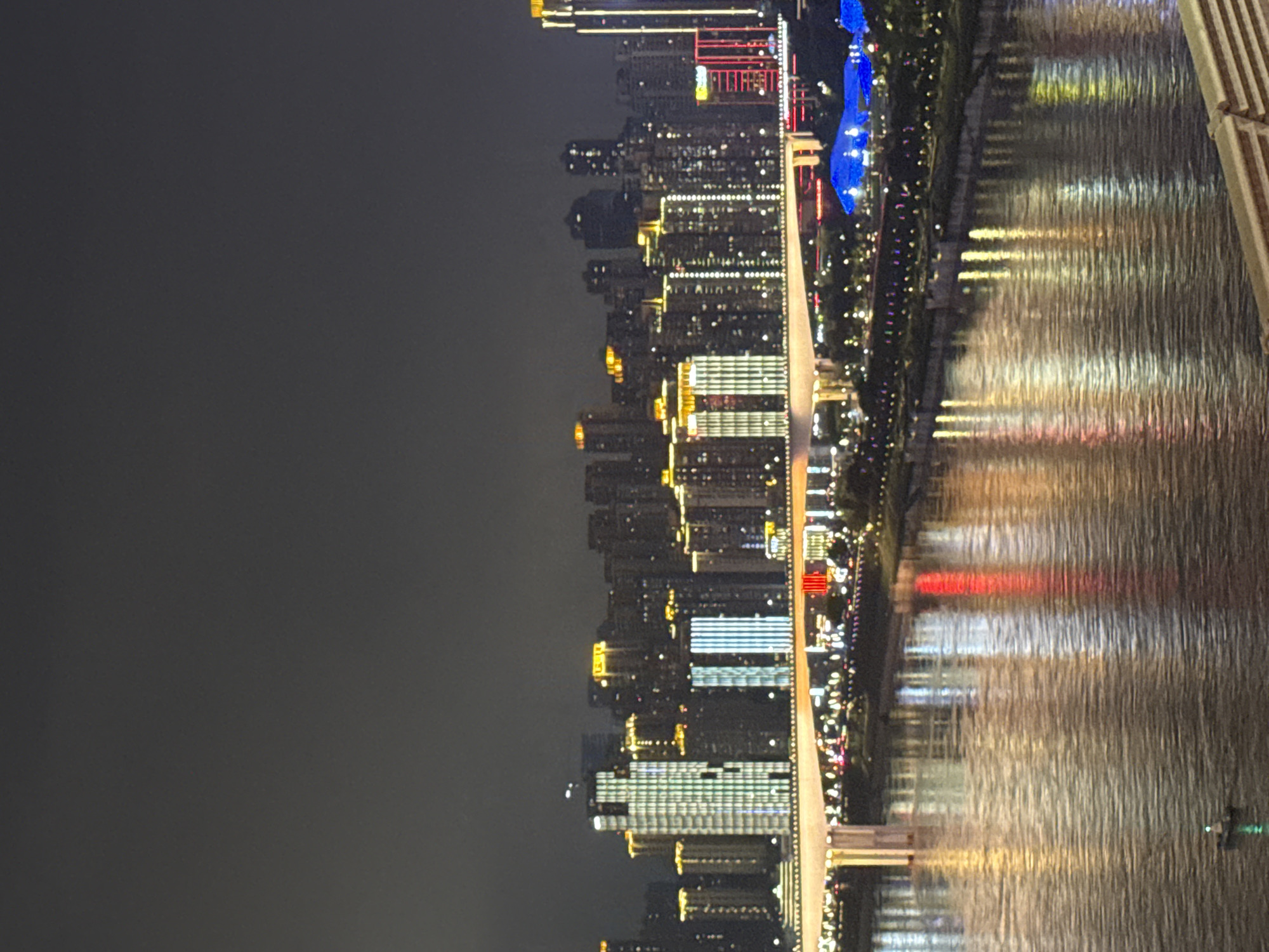 The image depicts a nighttime cityscape of Yuzhong District, China, dominated by a river and illuminated skyscrapers. A bridge with continuous lighting spans across the river, connecting the two sides of the city. The water of the river reflects the vibrant lights of the buildings and the bridge, creating a shimmering effect. In the foreground, a small boat with a green light is visible on the water, suggesting human activity. The background is filled with numerous high-rise buildings of varying heights and architectural styles, all lit up with a spectrum of colors, predominantly white, yellow, and green, with some red accents. The sky above is dark, indicating it is nighttime, with no visible celestial bodies. The overall scene portrays a bustling urban environment at night, characterized by modern architecture and the presence of water.