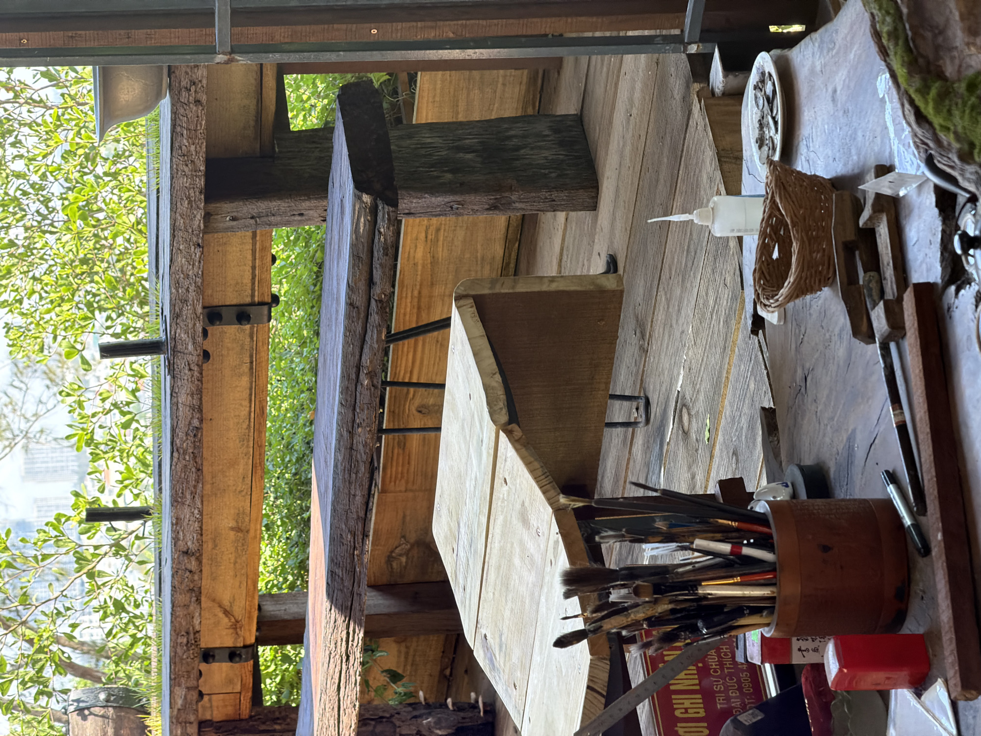 A sideways shot shows a workspace on a wooden deck in Đà Nẵng, Vietnam. A wooden table holds a container of paintbrushes, paint supplies, and other tools. A wooden chair sits beside the table. The space is open to a view of trees and a distant cityscape.