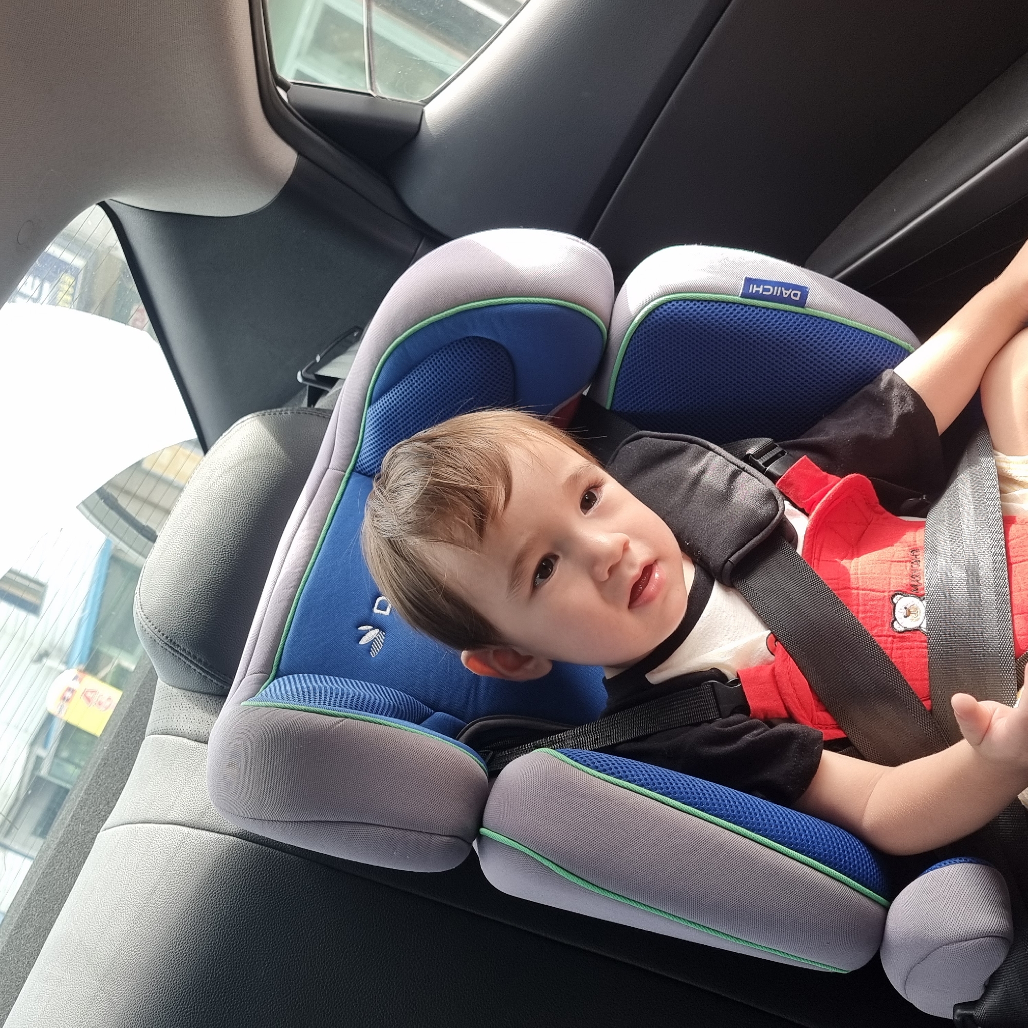A young child is sitting in a blue and gray car seat with a red harness. The child has light brown hair and is looking towards the camera. The car seat is positioned in the back seat of a car. The interior of the car is visible, including the black leather seats and the window. Outside the car, a blurred background shows buildings and a yellow sign. The image appears to have been taken on a sunny day.