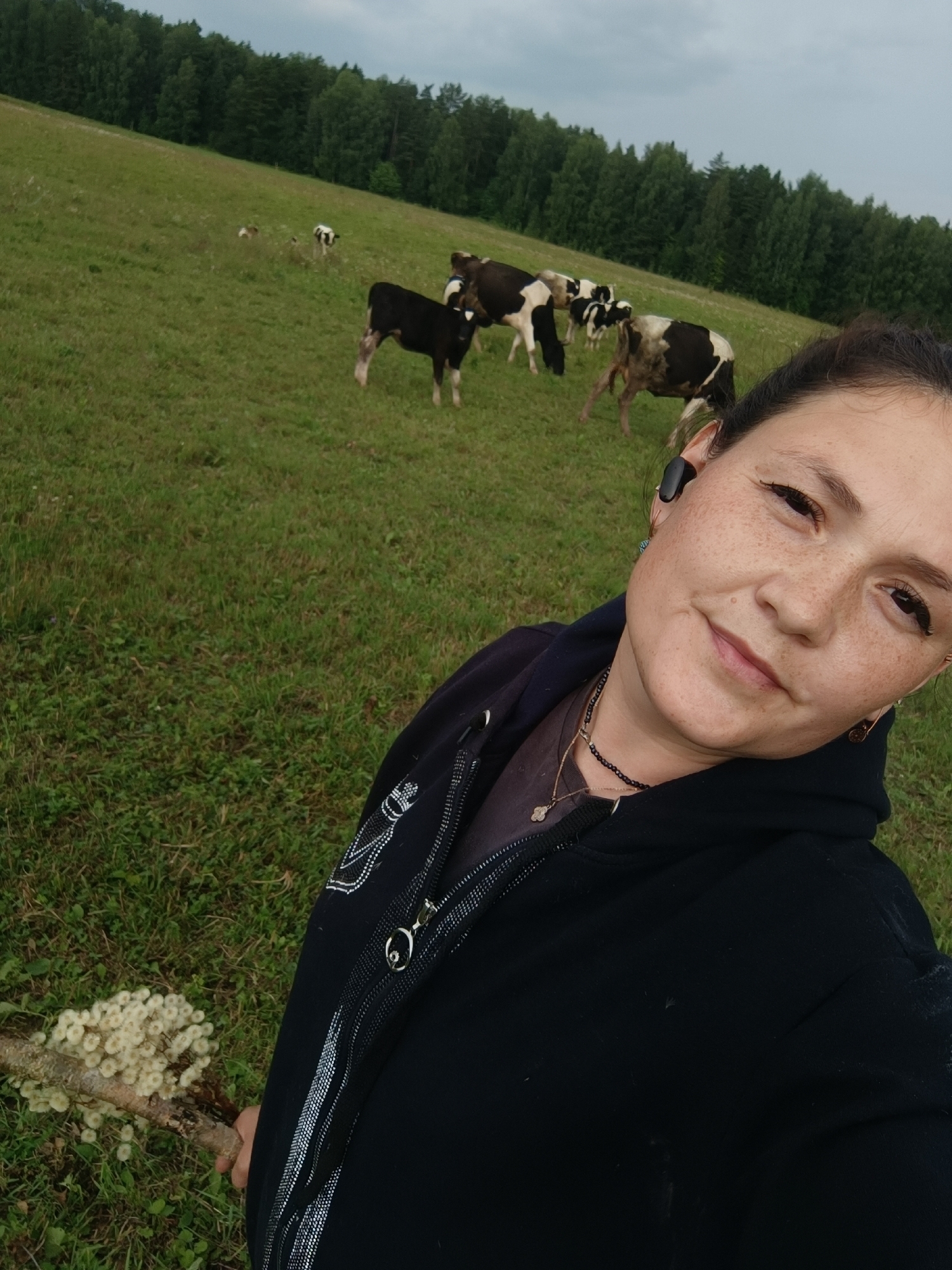 A woman is taking a selfie in a grassy field with cows in the background. She is wearing a black hoodie and has freckles on her face. A branch with small white flowers is visible in the foreground. The field is sloped and covered with green grass. In the background, there is a line of trees. The sky is overcast. There is no information that links this image to the city of Karsha, Russia.