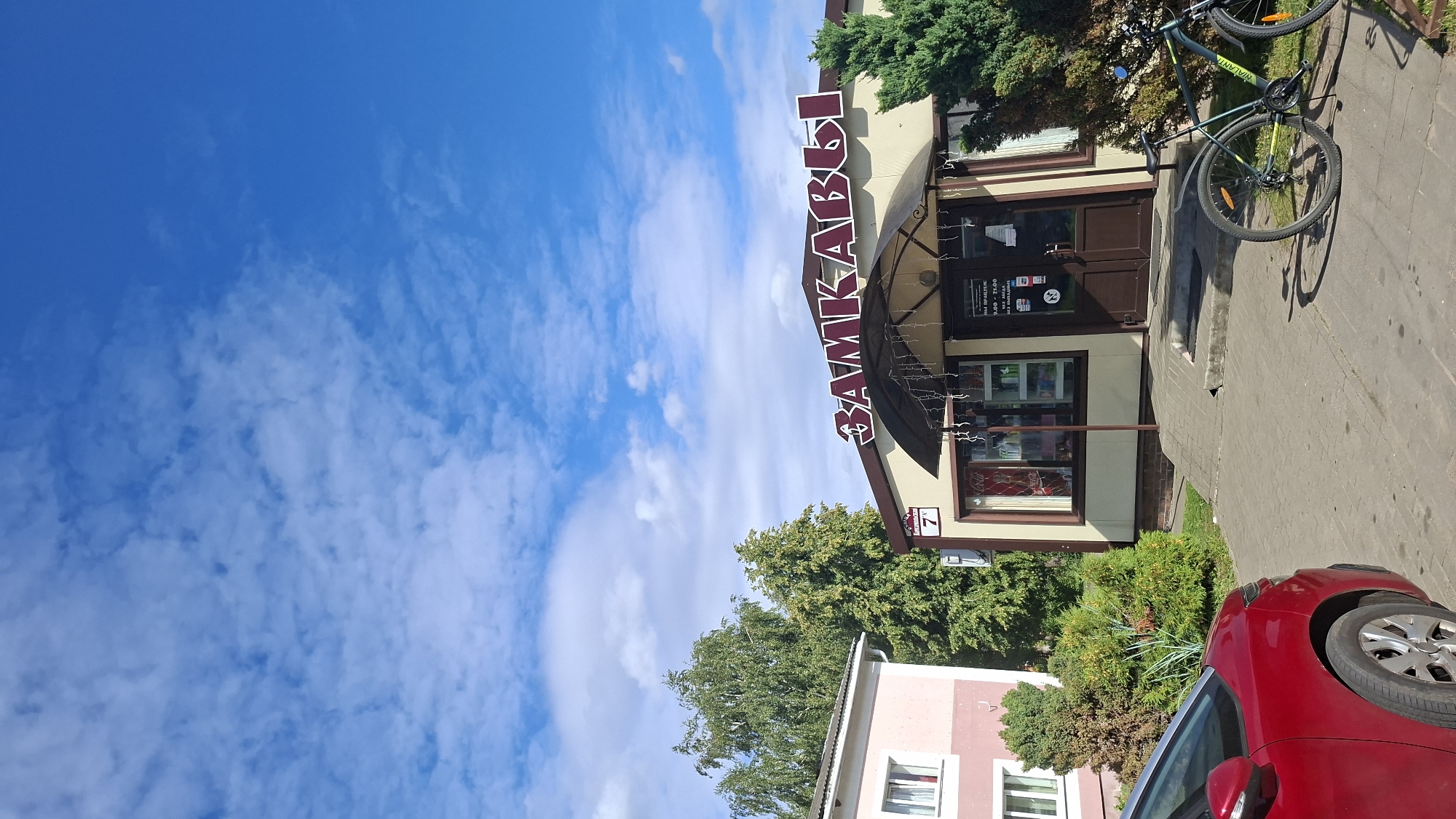 The image shows a store with a prominent sign reading "ЗАМКАВЫ" in large red letters. The store appears to be a retail establishment, possibly a food or convenience store, with items visible through the windows and a display shelf inside. Two doors are visible, one with a sign listing operating hours from 9:00 to 21:00. A bicycle is parked to the right of the entrance, and another bicycle is visible in the lower right corner of the image. A red car is parked to the left, partially obscuring the foreground. To the left of the store is a light pink building with windows. Green trees and bushes surround the store and the adjacent building. The sky is blue with scattered white clouds. The location is identified as Myadzyel, Belarus.