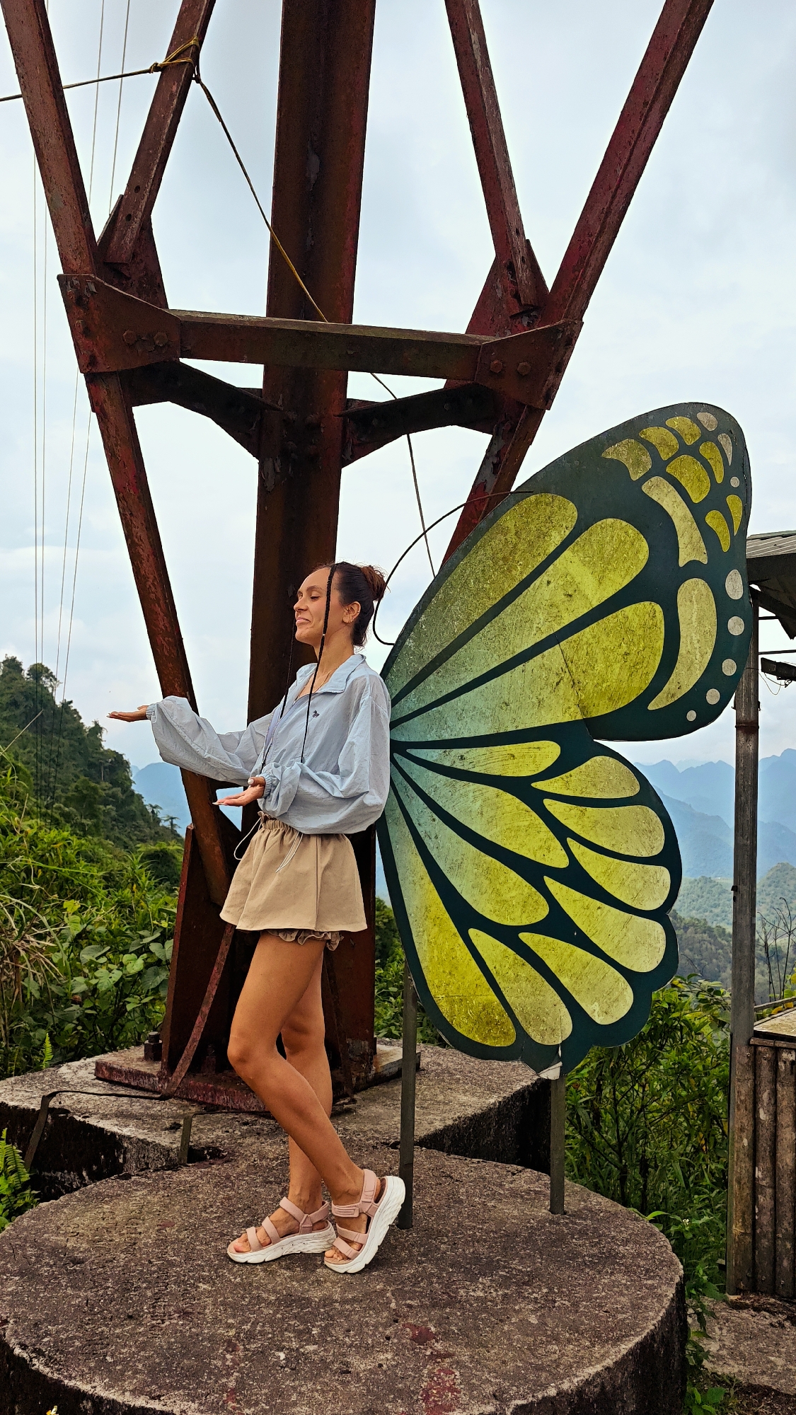 A full shot captures a woman standing on a concrete platform in Tam Son, Vietnam. She poses next to a decorative butterfly wing display, utilizing it as a prop for her photo. The wings are painted in shades of yellow and dark green with white circles along the edges. The woman is clad in a light blue, button-up blouse, and a khaki-colored skirt. She wears pink and white sandals and has her hair styled in a bun. She has her eyes closed, and a content expression on her face.
Behind the woman, a large, rusty metal structure rises toward the sky. The structure is an antenna tower. The surrounding environment is lush with greenery, and mountains can be seen in the distance. The sky is overcast, casting a soft, diffused light over the scene.