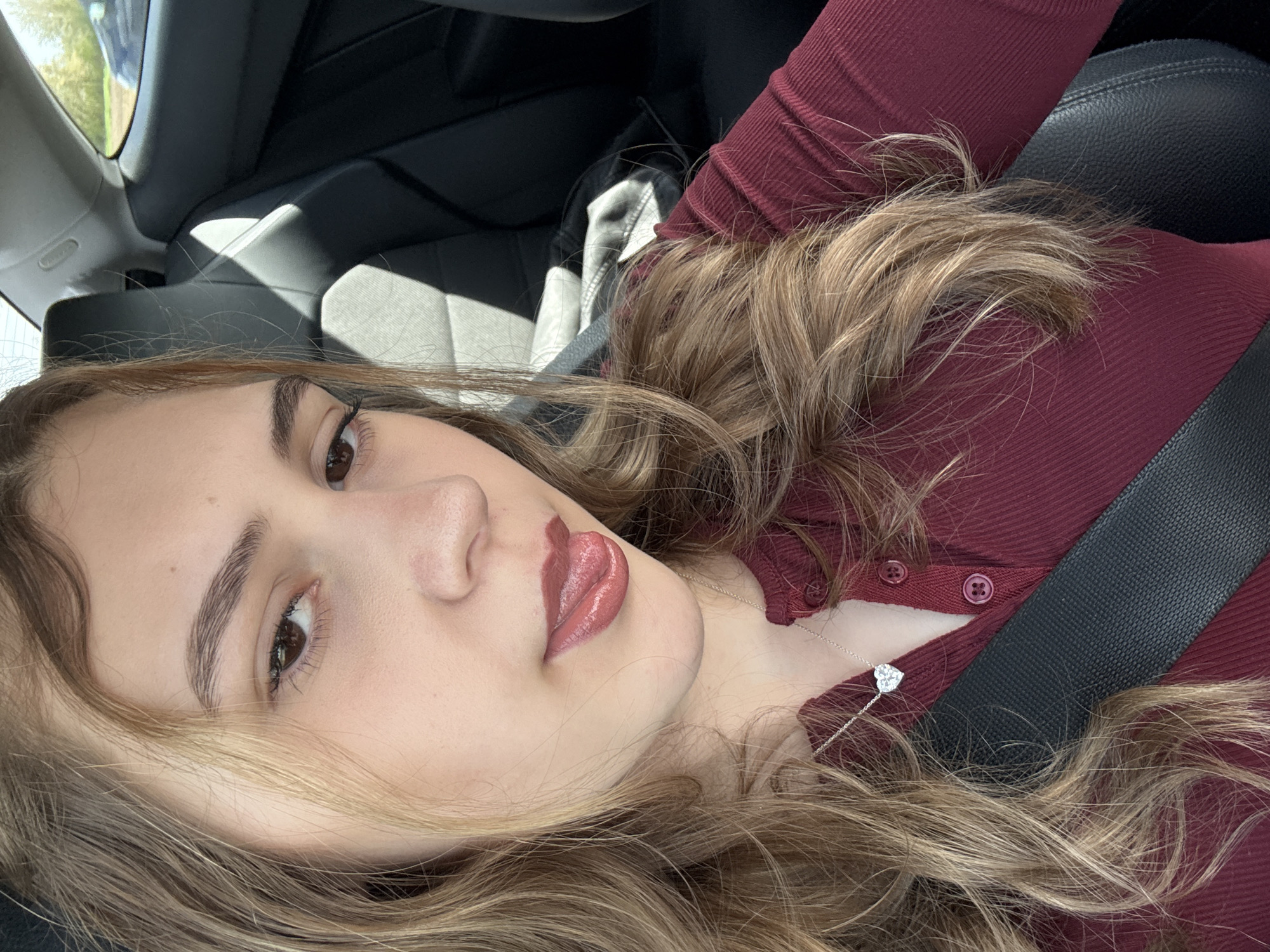 A young woman with long, wavy blonde hair lies in a car wearing a maroon, ribbed, button-up shirt and a seatbelt. She has fair skin, brown eyes, and is wearing lipstick. A silver necklace with a heart pendant is visible. The car's interior is black leather. The location in the city of Herrlisheim, France is not discernible from the image.