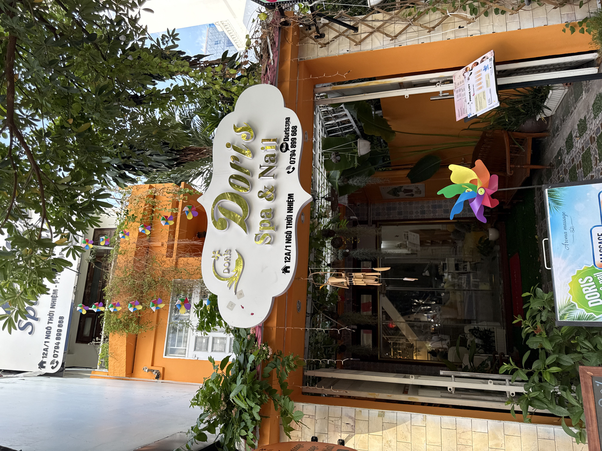 The image shows the exterior of "Doris Spa & Nail" located at 12A/1 Ngo Thoi Nhiem Street in Nha Trang, Vietnam. The building features an orange facade with a white, cloud-shaped sign at the entrance. The sign displays the spa's name in gold lettering, "Doris Spa & Nail," along with an address and phone number: 0794 899 668.
Below the sign, the entrance to the spa is visible, with glass doors and windows revealing the interior. Hanging from the eaves are decorative items, including colorful pinwheels and a wind chime. Lush green plants cascade down the building and surround the entrance. A wooden bench is placed to the left of the entrance, and a price list is displayed on a stand to the right. The street in front of the spa has a tiled walkway. The overall scene is bright and inviting, with a focus on natural elements and a spa-like atmosphere. No people are prominently visible in the image.