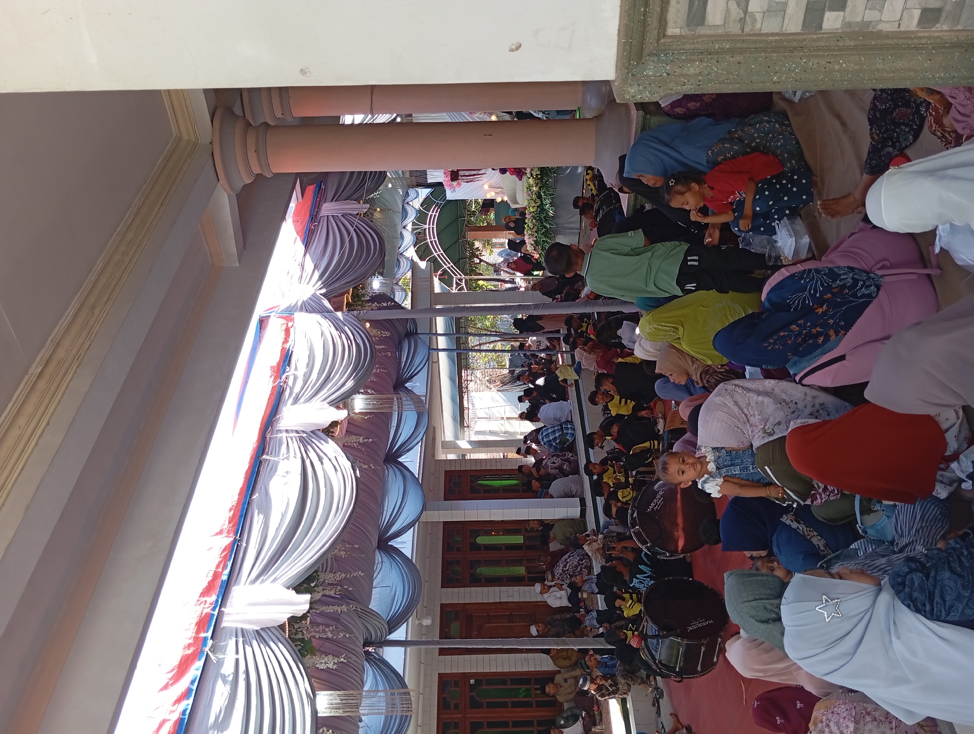 The image depicts an outdoor gathering in a semi-covered area, likely a community event or celebration, in Padurungan, Indonesia. A large tent with draped fabric, featuring blue, white, and red accents, provides shade. Numerous people, including men, women, and children, are seated on the ground on mats. Some are dressed in traditional Islamic attire, such as hijabs and koko shirts.
In the foreground, a young child in a blue denim outfit stands out, looking towards the camera. Several women are seated around her, also wearing hijabs in various colors. To the left, two large bass drums are visible, suggesting a musical or ceremonial aspect to the event. In the background, a raised platform decorated with flowers and white fabric can be seen, possibly a stage or focal point for the gathering. The overall environment appears to be a public space, with tiled walls and architectural columns indicating a building or structure nearby. The lighting suggests it is daytime.