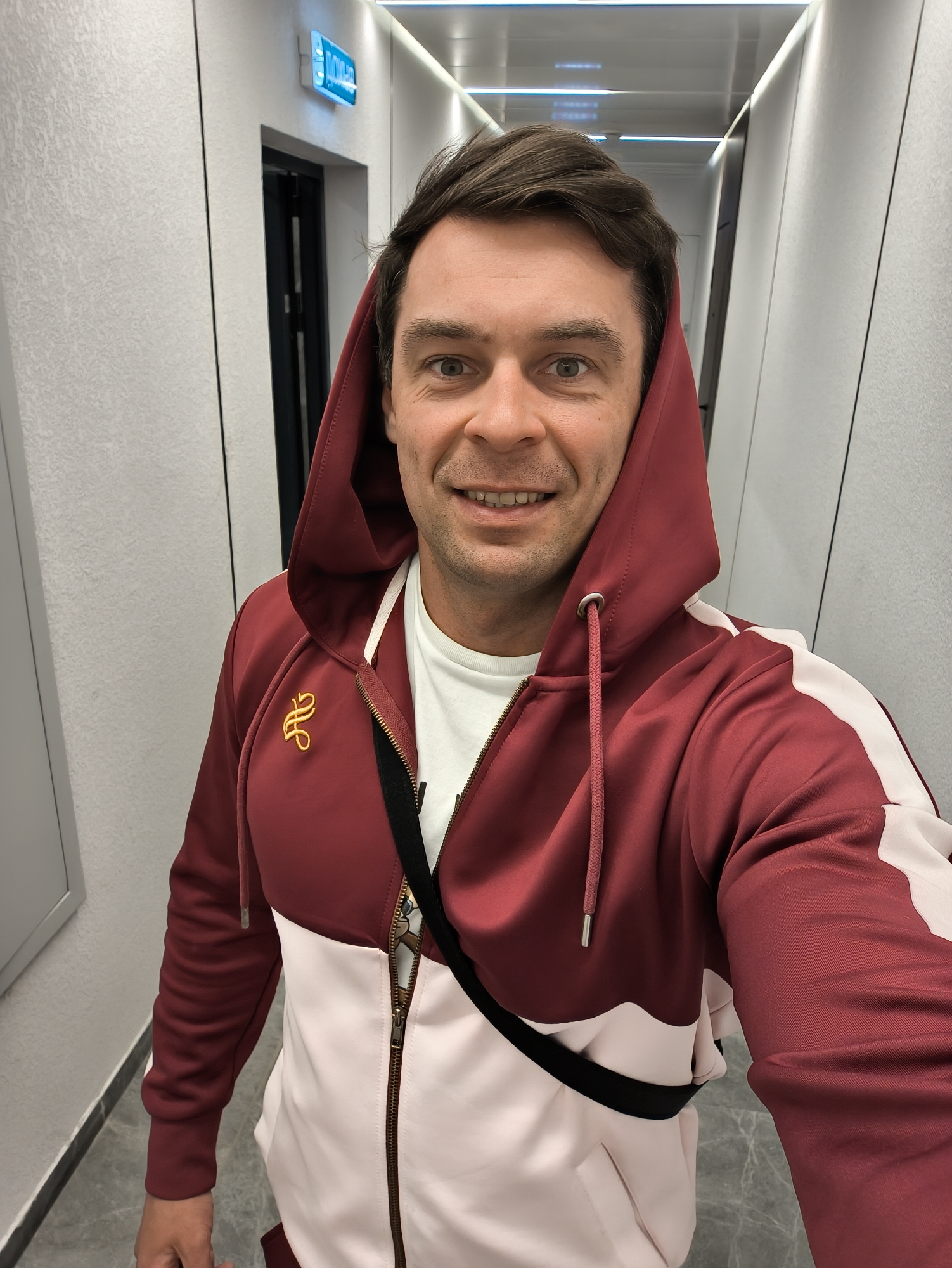 A cheerful man takes a selfie in a brightly lit, modern hallway. He is fair-skinned with short dark hair and brown eyes, smiling broadly at the camera, revealing his teeth. He wears a two-toned zippered hoodie; the upper section and sleeves are a rich maroon, while the lower body and parts of the sleeves are light pink or off-white. A golden, stylized emblem, resembling intertwined letters, is visible on the left chest of his hoodie. Underneath, a white t-shirt can be seen, and a black strap, possibly from a bag, crosses his chest. The hoodie features maroon drawstrings with silver tips and a gold-colored zipper.
The setting is an indoor corridor with light gray, textured walls and a gray tiled floor. The white ceiling is equipped with linear strip lights, providing ample illumination. A dark-colored door with a matching frame is partially visible on the left side of the hallway. Above this door, a blue light fixture displays glowing Cyrillic text that clearly reads "МОСКВА" (MOSKVA), followed by what appears to be "14" or "24", indicating the location as Moscow, Russia. The man's expression is happy and friendly.