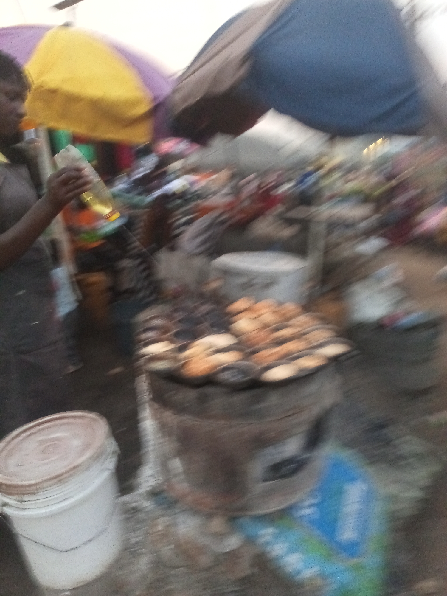 The image depicts a street scene in Rafin Par, Nigeria, with a blurred, low-resolution quality that makes precise identification challenging. In the foreground, a white bucket with a metallic handle is visible on the left. To the right of the bucket, a cylindrical metal grill or oven is present, with rows of round, light-brown food items, possibly pastries or small breads, cooking on it. Smoke rises from the grill, indicating it is in use.
A person, whose face is obscured by blur and shadow, is standing to the left of the grill, holding a clear bottle containing a yellowish liquid. The person appears to be wearing a dark-colored garment.
Behind the grill and the person, there are glimpses of market stalls with colorful umbrellas, one purple and yellow, and another large blue one. The background is filled with a chaotic blur of colors and shapes, suggesting a crowded marketplace environment with more stalls and people. The ground appears to be uneven and dusty. A blue and white object with some text, possibly a plastic bag or a sign, is visible on the ground to the right of the grill. The overall impression is of an outdoor market in a bustling urban or semi-urban setting.