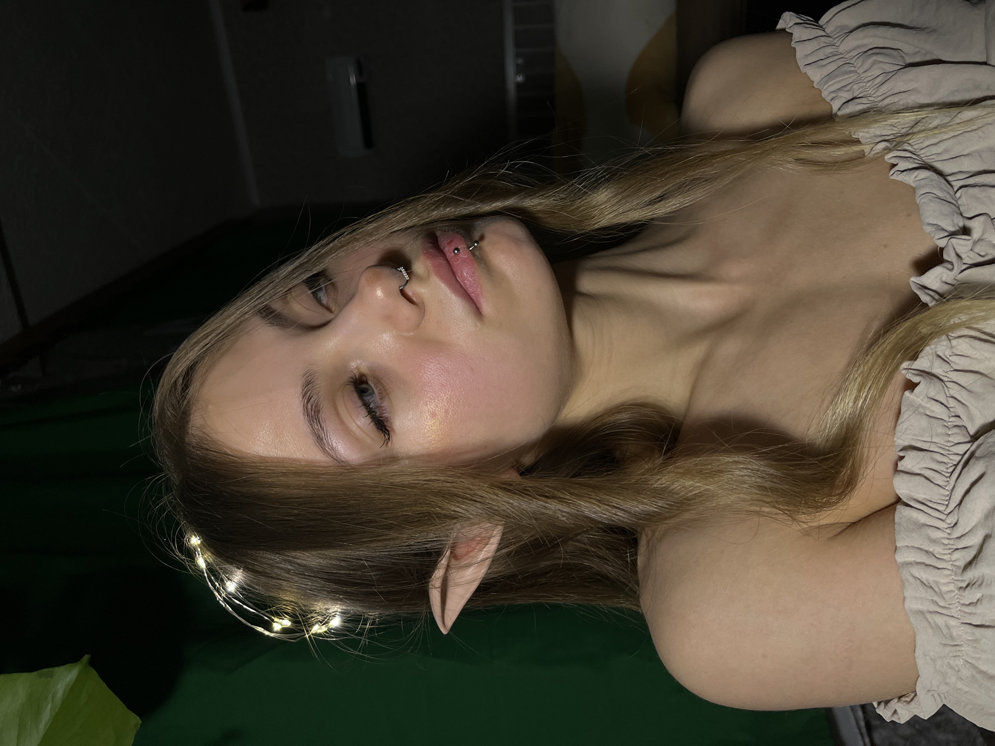 A young, fair-skinned person with blonde hair lies on a green surface, partially obscured by what appears to be a plant. The individual is adorned with a string of small lights in their hair and has pointed ears, possibly part of a costume. The person has lip and nose piercings. They are wearing a beige, ruffled top. The location in the city of Фоки, Russia, cannot be determined from the image.