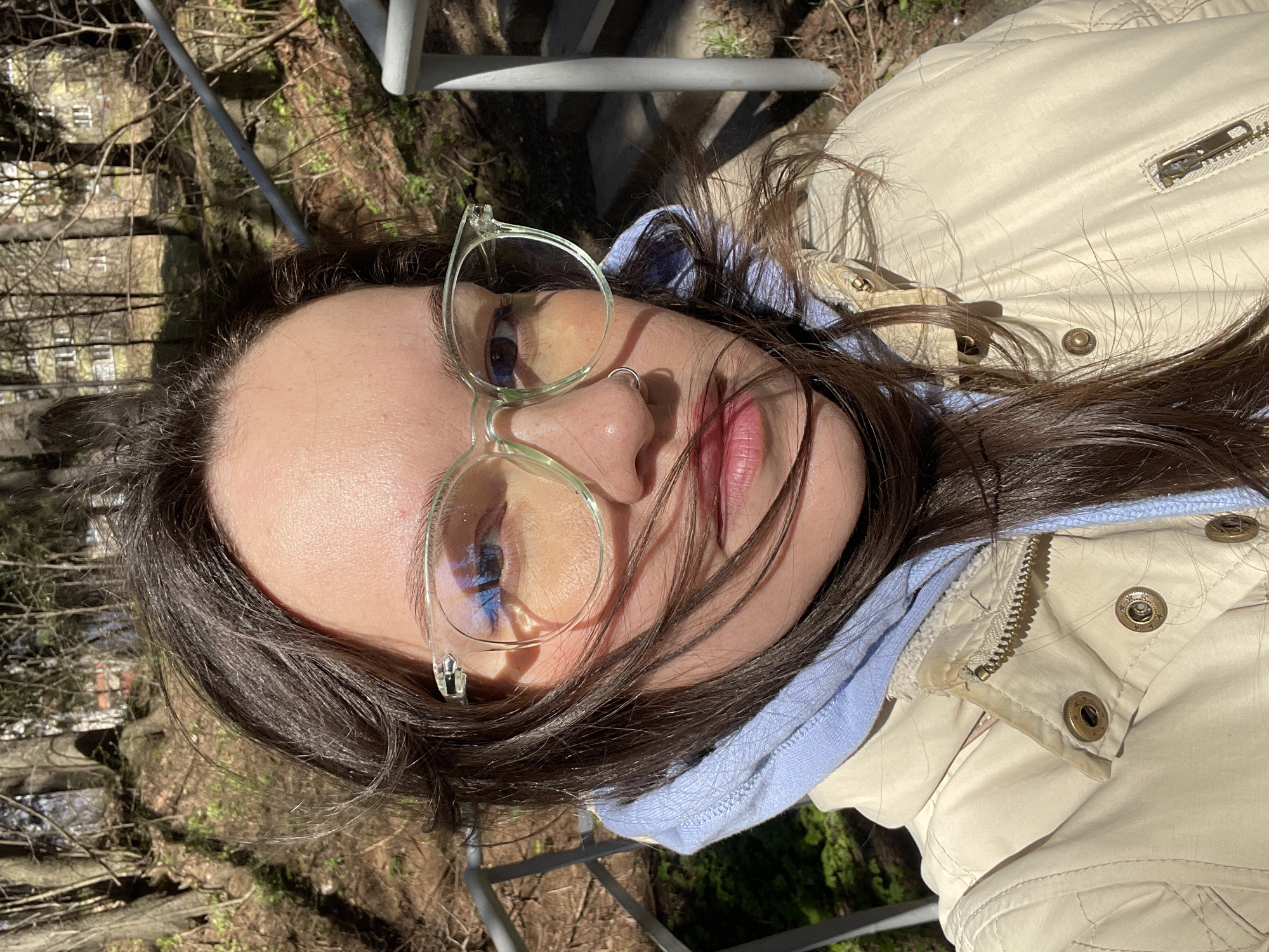 This media file features a close-up portrait of a person, likely a young adult female, with their head tilted. They have long, dark brown hair, which is somewhat dishevelled and falls across their face and shoulders. The person is wearing light green-rimmed glasses, through which reflections of a bright blue sky and clouds are visible in the lenses. A subtle nose ring adorns their left nostril, and their lips are slightly parted, revealing a natural pink hue.
The individual is dressed in a casual layered outfit: a light blue hooded sweatshirt underneath a light beige or cream-colored quilted jacket. The jacket features metallic buttons and a zipper, with some visible wear around the collar area.
The scene is set outdoors on a bright, sunny day, evidenced by the strong lighting and distinct shadows cast by hair strands onto the jacket. The background, though out of focus, reveals an elevated vantage point, possibly a balcony or bridge, with metal railings. Beyond this, the natural environment features numerous bare tree branches and sparse ground cover, suggesting a season of late autumn or early spring. In the distance, through the branches, several older-looking buildings with multiple windows are visible, alongside what appears to be a reflective body of water, possibly a river or stream. The overall mood is calm and contemplative. No discernible text is visible in the image. The location context for this scene is Izhevsk, Russia.