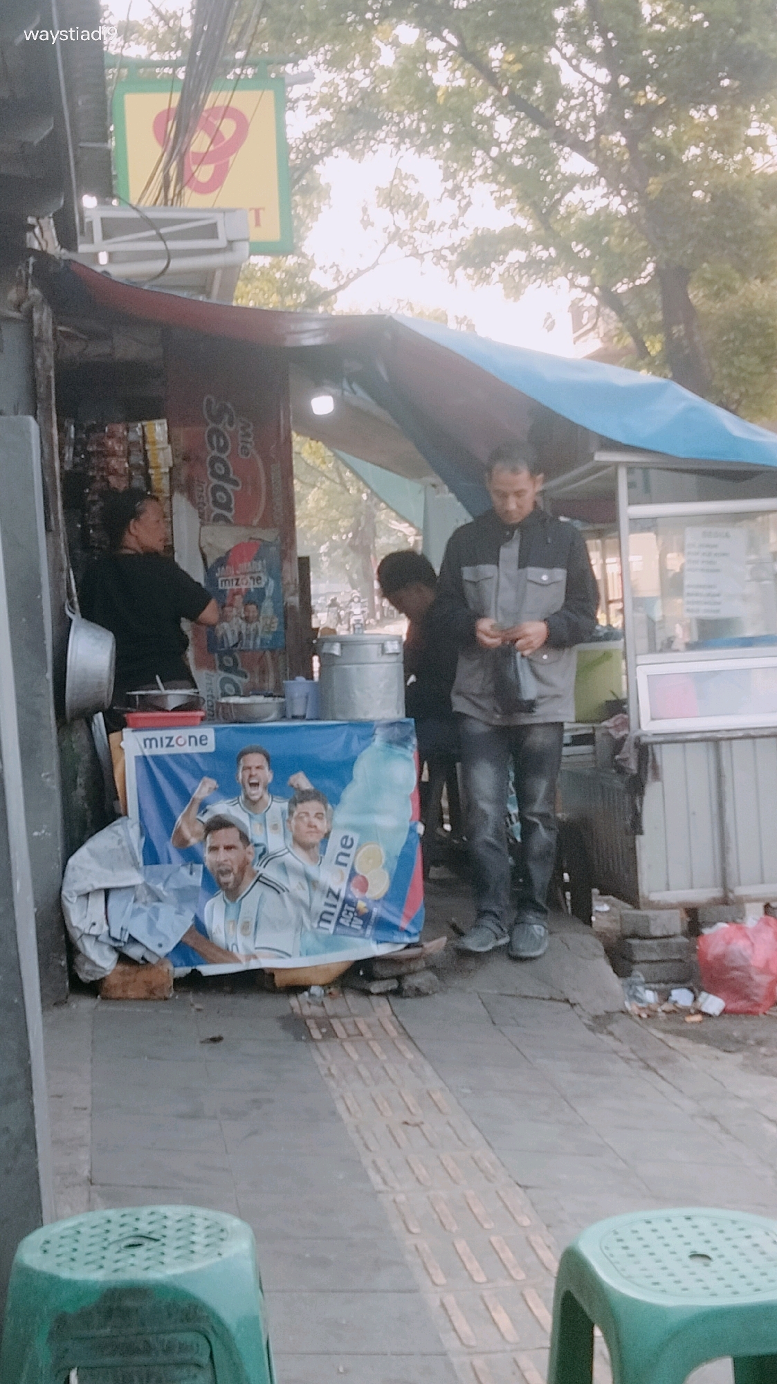 An outdoor food stall is set up on a sidewalk in a city. A man in a grey and black jacket and dark jeans stands in front of the stall, looking at his phone. Behind him, a woman with dark hair tied back is serving customers. A large blue banner with images of soccer players and a bottle of Mizone drink is displayed at the front of the stall. A yellow sign with a red infinity symbol is visible above the stall, and a street sign with "waystiadi9" is on the upper left of the image. The ground is paved with tiles and a textured strip for visually impaired pedestrians. In the foreground, a green plastic stool is visible. Trash is scattered on the right side of the image. The scene appears to be in Bandung City, Indonesia, suggested by the text "waystiadi9" which might be a street name or identifier.
