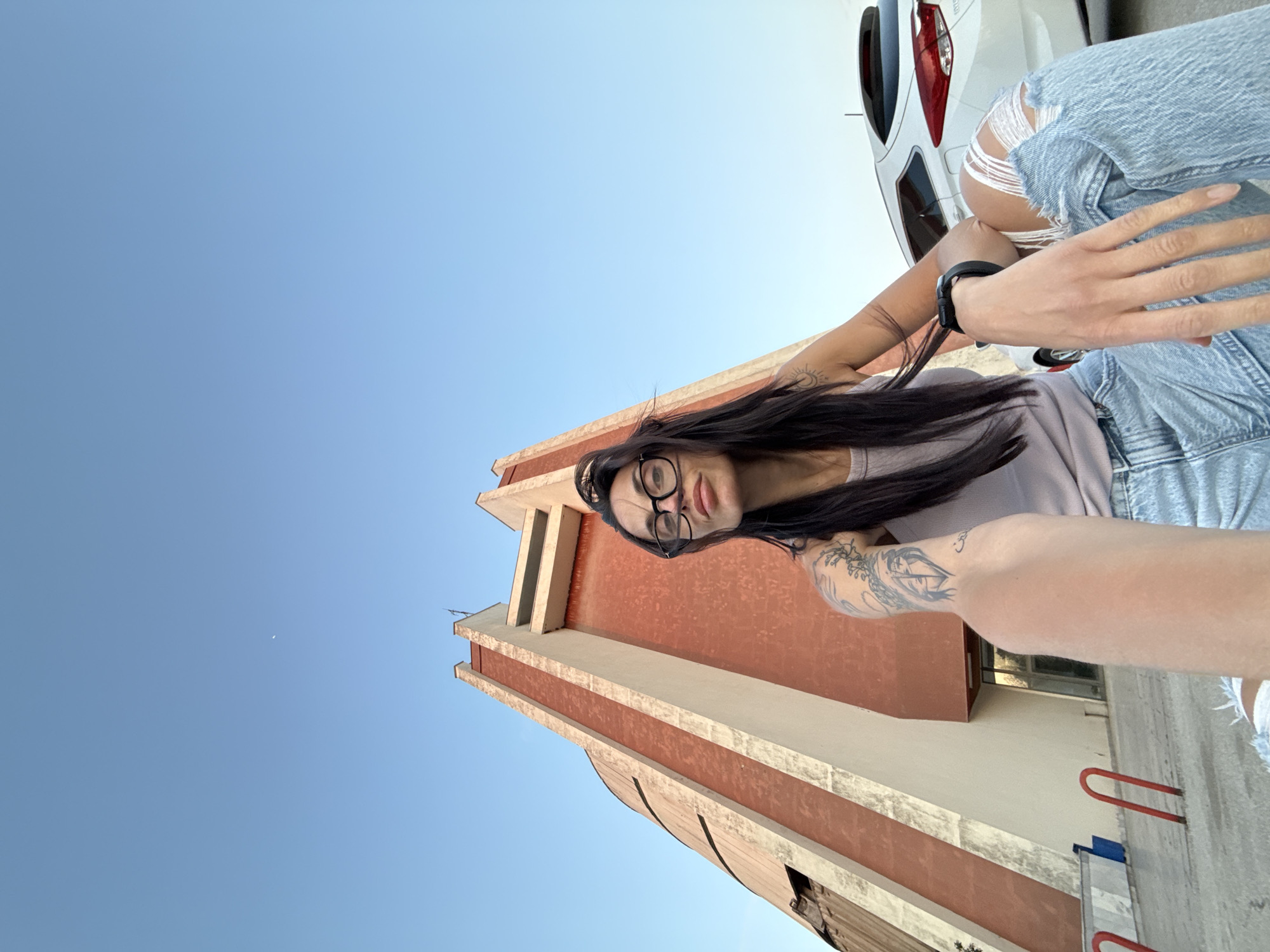 A woman with dark hair and glasses is posing for a selfie in Pleven, Bulgaria. She is standing in front of the Church of Saint Nicholas. A white car is visible in the background. The sky is clear and blue.