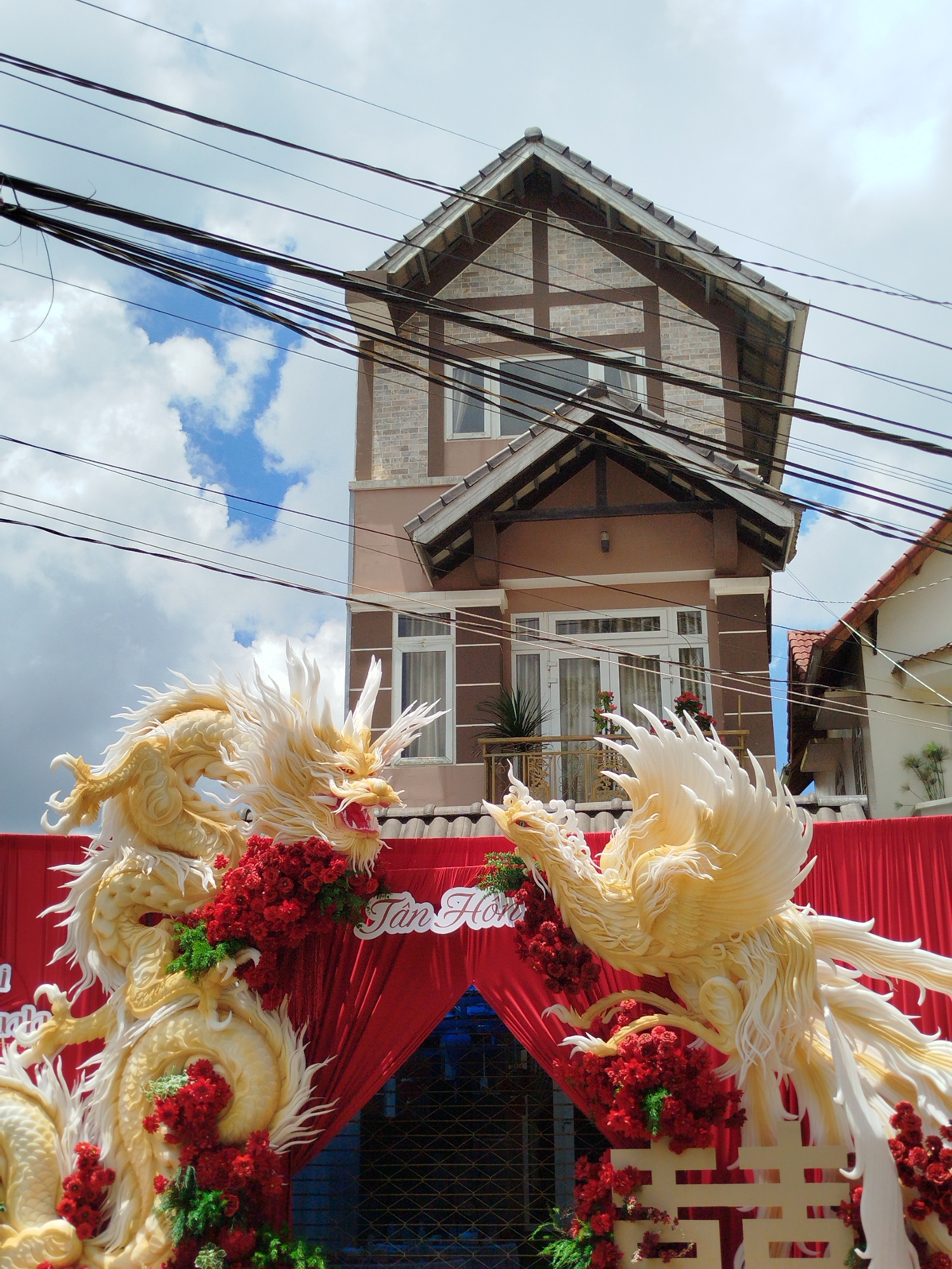 In the city of Xuân Trường - Đà Lạt, Vietnam, a wedding archway is decorated with a large, ornate dragon and phoenix sculpture made of white and gold material. The sculptures are intertwined with red flowers and greenery. The archway is draped with red fabric, and the words "Tân Hôn" (meaning "New Marriage" in Vietnamese) are written in white cursive script on the fabric. In the background, a multi-story house with brown and tan brick siding and a dark roof is visible, along with a section of another building. Overhead, a tangle of dark power lines crisscross the bright blue sky, which is dotted with white clouds.