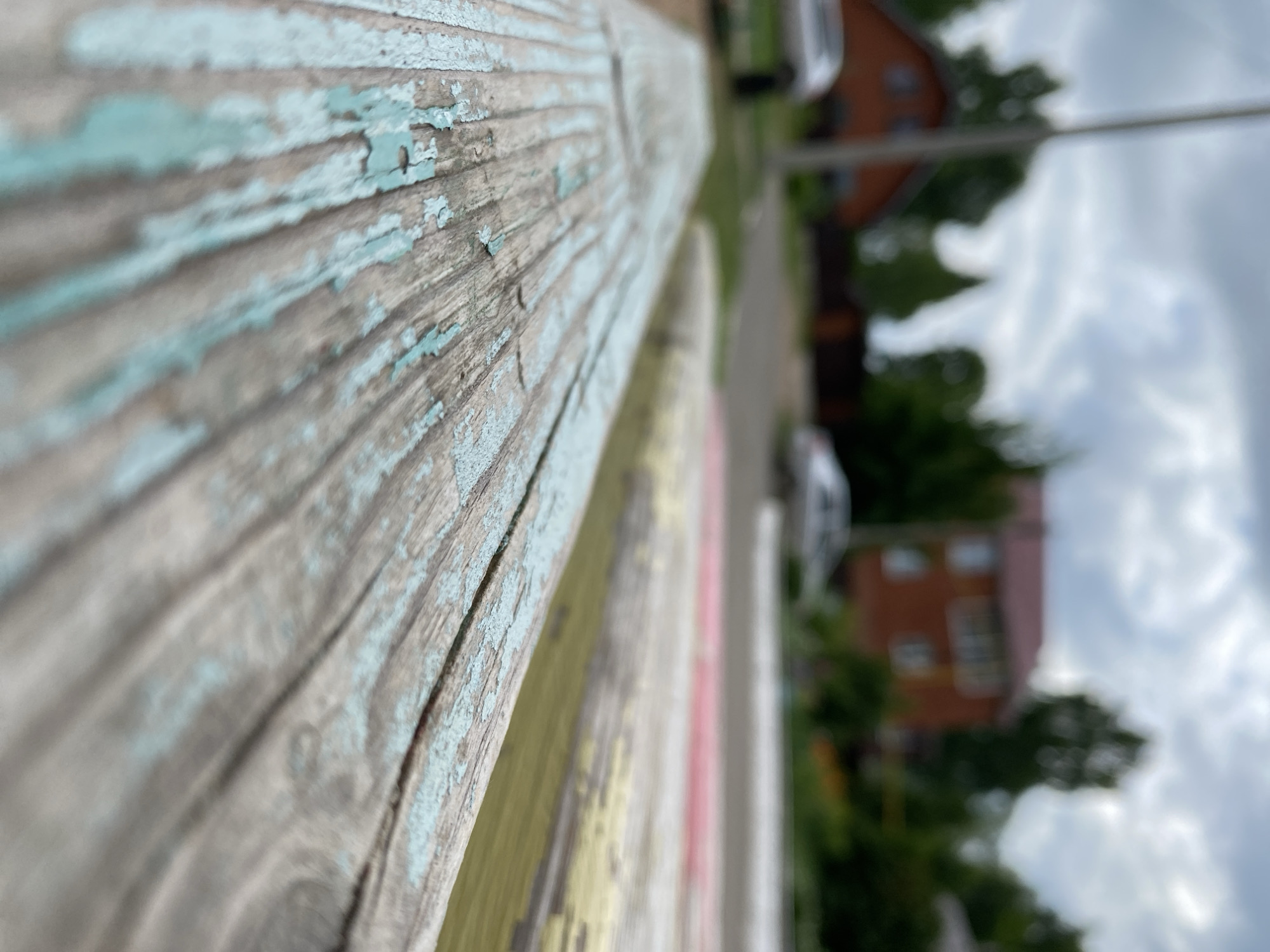 The image shows a close-up of a weathered, light blue wooden surface, likely a bench, with peeling paint. In the blurred background, a red brick house with multiple windows and a dark roof is visible. Several green trees surround the house. A silver car is parked in front of the house, and another silver car is partially visible on the right side of the frame. The sky is overcast with grey clouds. The scene appears to be in a residential area, potentially a dacha or a village, in Votkinsk, Russia.