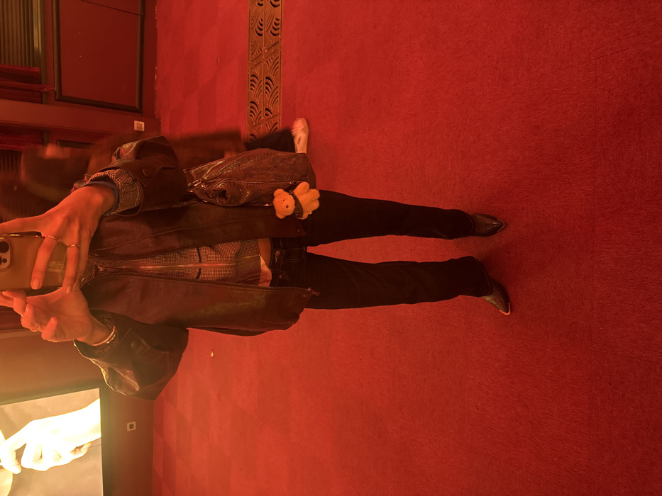 A person stands on red carpet, taking a selfie with a phone. They wear black pants and brown leather shoes. A brown leather jacket covers a patterned shirt. A brown leather purse hangs over their shoulder with a small bear attached. Red walls surround the carpeted area. A glass window with blurred images is visible to the left.