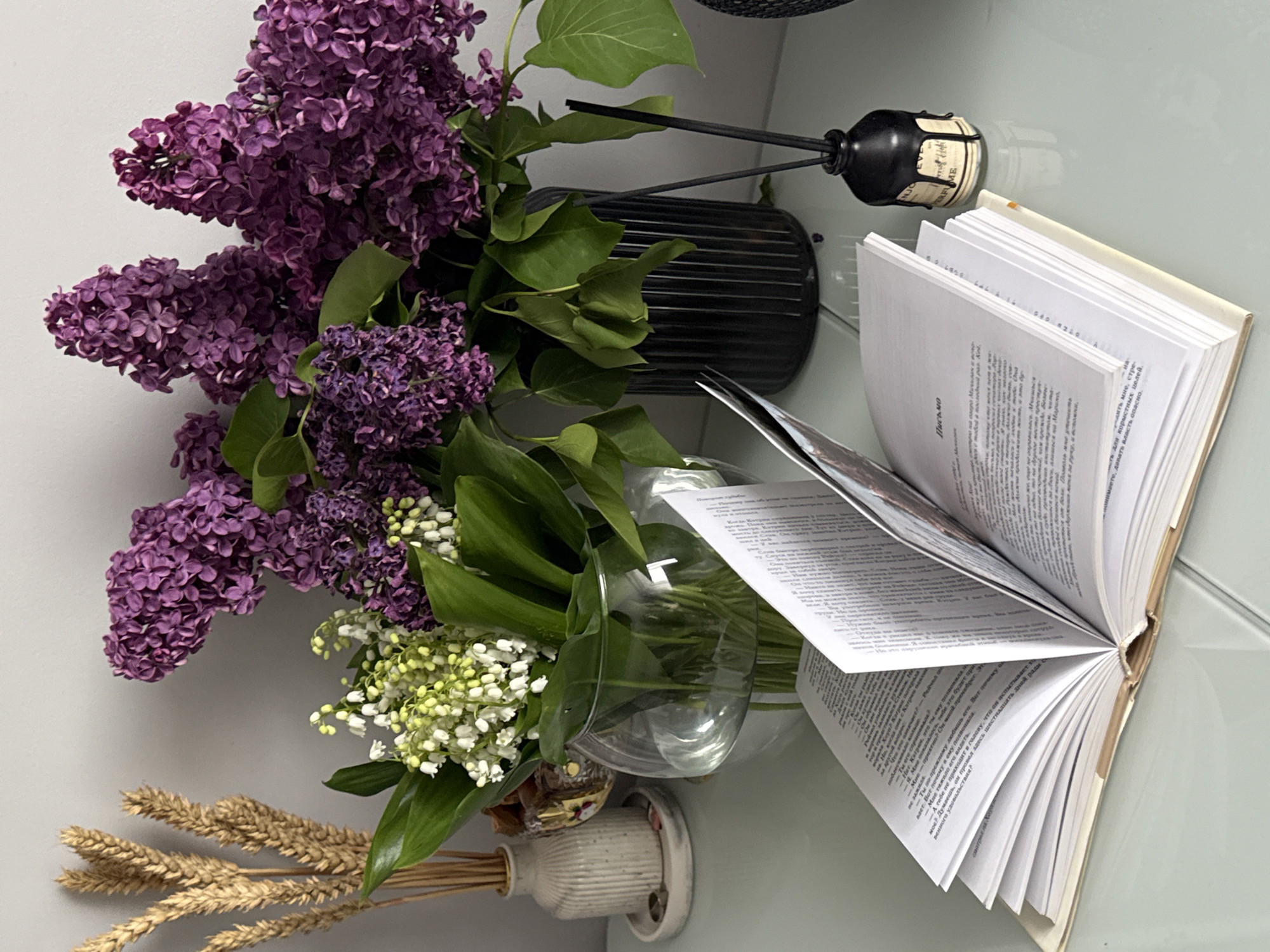 An indoor still life arrangement is captured from a slightly elevated perspective, showcasing a collection of items on a light-colored, reflective surface. The scene is dominated by floral elements, including a lush bouquet of vibrant purple lilacs with their green leaves, alongside delicate white lily-of-the-valley flowers. These are displayed in clear glass vases, with the lilacs in a larger, dark ribbed vase (partially visible) and the lily-of-the-valley in a smaller clear vase.
To the right of the flowers, an open book with numerous white pages lies spread, revealing text printed in Cyrillic script. A black reed diffuser with reeds stands in the background, its dark bottle featuring a label with partial English text reading "NJO" and "ERFUME" (likely from "PERFUME"). In the bottom left, a small white, ribbed ceramic vase holds several stalks of dried golden wheat.
The activity suggested is contemplation or reading, indicated by the open book. The fresh flowers imply a springtime setting. The visible text in the book includes: "Письмо" (Letter) as a heading, and phrases such as "поблескивало", "Ты его спрашиваешь", "Мне понятно?", "не зажила", "Мне тяжело его видеть", "Ты по-прежнему любишь его", "A разве не приходит в голову, что он испытывает не самого великого удовольствия?". On the right page, fragments like "мне, стре-", "для корыстных целей.", and "давать власть опасно." are legible. The overall mood is tranquil and domestic.