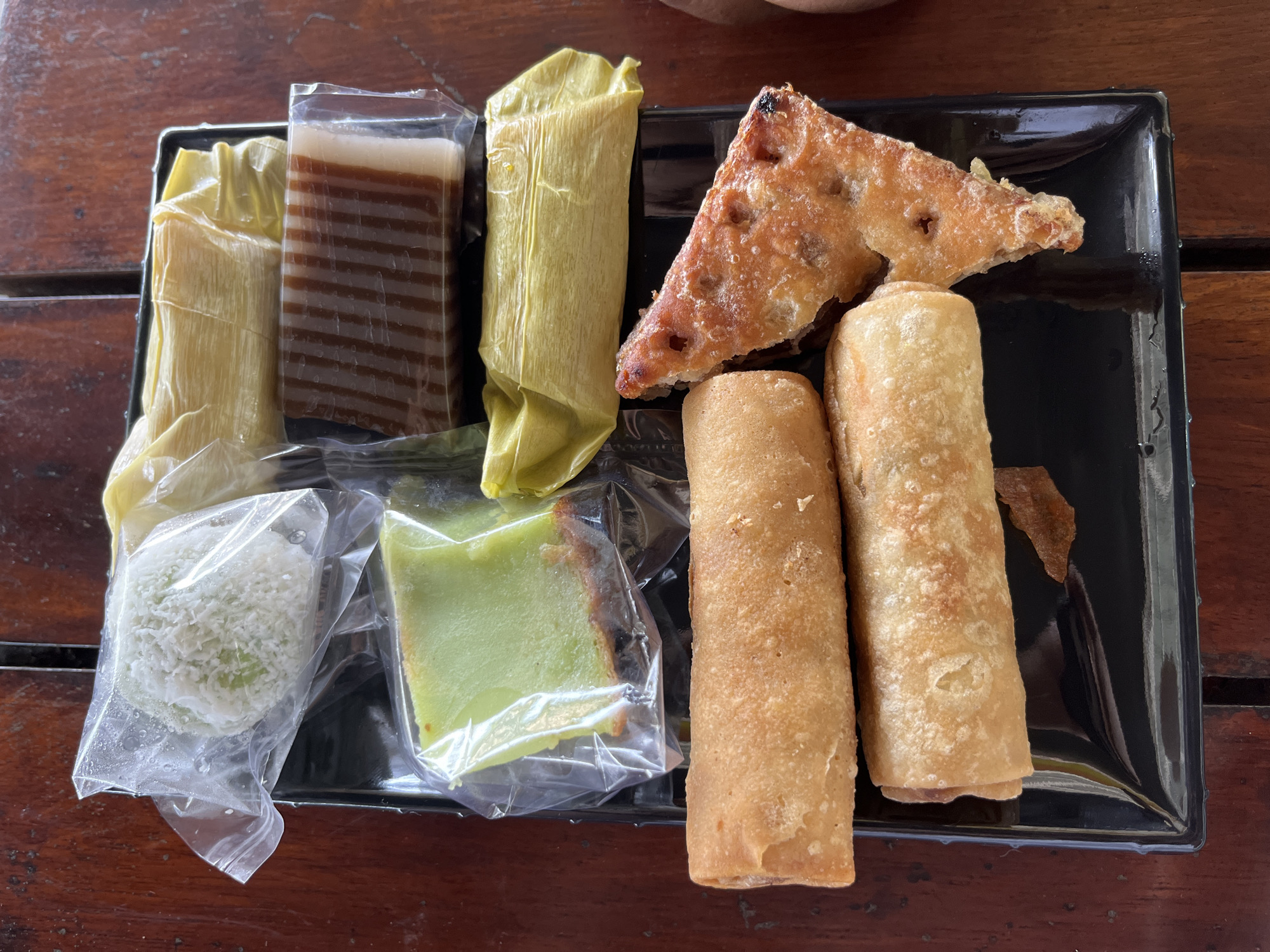 A top-down shot showcases a variety of Indonesian traditional snacks, commonly known as 'kueh', neatly arranged on a black rectangular serving tray. The tray rests on a rustic wooden table, displaying its visible grain.
The assortment of snacks includes:
- A rectangular, multi-layered dessert with alternating brown and white stripes, likely a 'kue lapis', sealed in clear plastic packaging.
- Two elongated items, possibly steamed rice cakes or dumplings, wrapped in light yellow, pliable leaves, resembling banana leaves or corn husks.
- A triangular, golden-brown fried pastry, possibly a sweet fritter like 'pisang goreng' (fried banana) or a savory equivalent, exhibiting a crispy texture.
- Two cylindrical, golden-brown rolled items, resembling spring rolls ('lumpia') or sweet crepes ('dadar gulung').
- A small, roundish green snack, possibly 'klepon' or 'onde-onde', generously coated in grated white coconut and enclosed in a clear plastic bag.
- A square slice of green cake, likely pandan-flavored 'kue talam' or a similar dessert, with a slightly caramelized or browned edge, also packaged in clear plastic.
All items appear to be individually packaged, suggesting they are either for takeaway, sale, or to maintain freshness. The image captures a diverse selection of local Indonesian delicacies from Meunasahaluedrienlhoksukon Barat, Indonesia, offering a range of textures and flavors. No text is visible in the image.