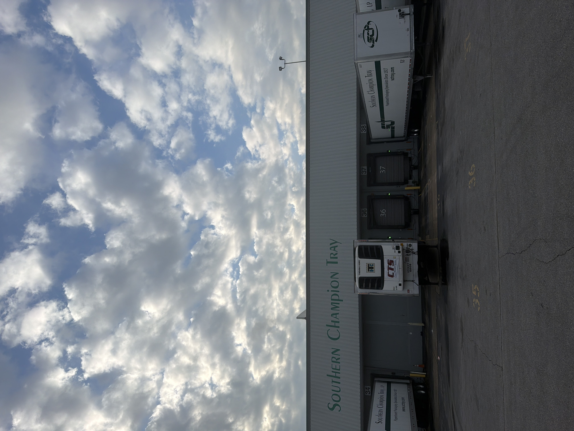 The image captures a loading dock area of a commercial building with "SOUTHERN CHAMPION TRAY" prominently displayed on its facade. Several semi-trailers are parked at the loading bays, identified by numbers 34, 36, and 37. One trailer displays the text "SOUTHERN CHAMPION TRAY, LP" and a website address "sctray.com". Another trailer has a "CTS" logo and "THERMO KING" branding, indicating it's a refrigerated unit. The environment appears to be an industrial or logistics facility. The sky above is partly cloudy with dramatic cloud formations. The ground is asphalt, with painted numbers "35", "36", and "37" visible in yellow. The scene suggests a business operation involving shipping and receiving. This facility is located in Chattanooga, United States of America.