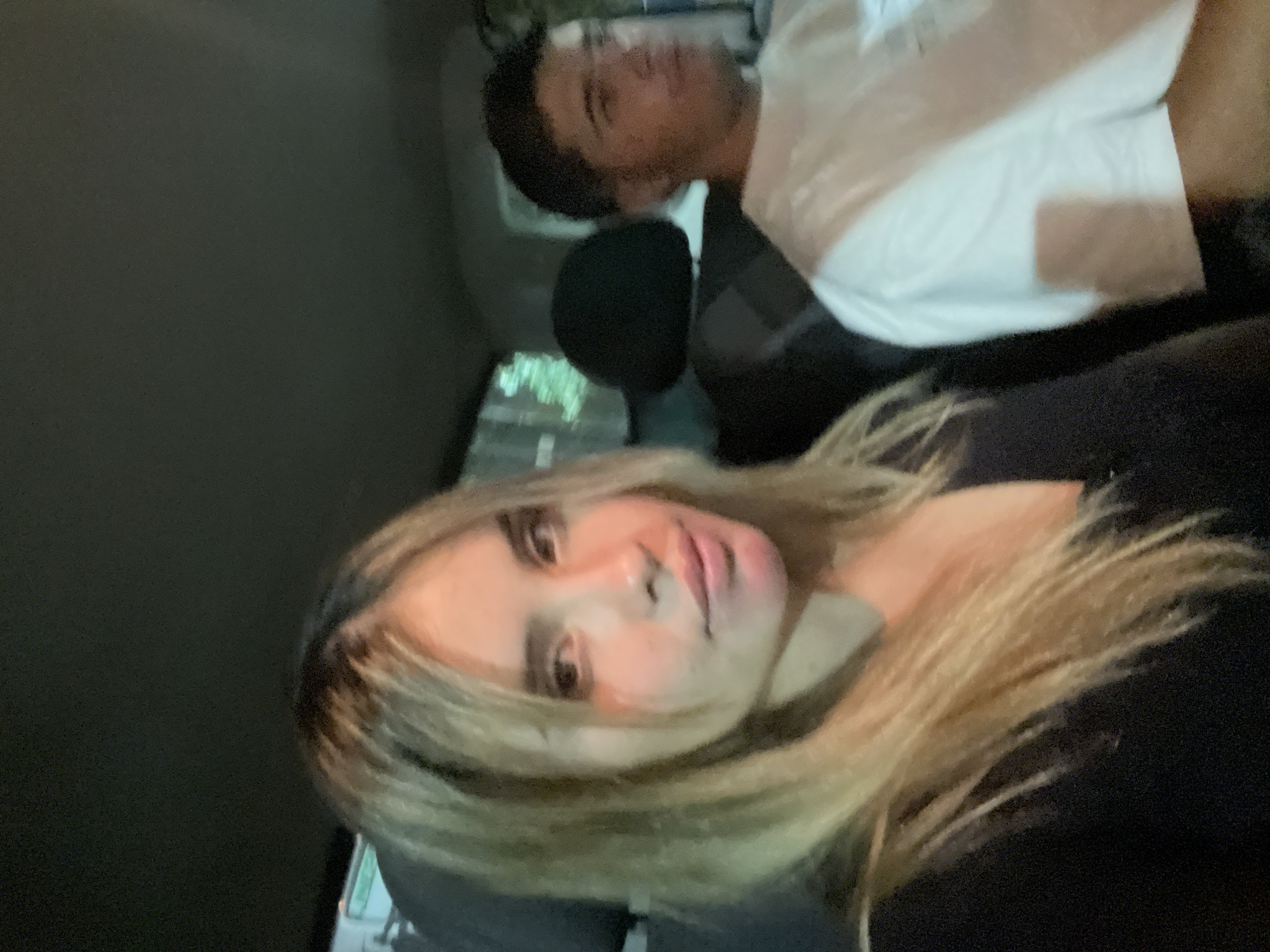 Two individuals are present within a vehicle. The foreground subject is a female with blonde hair wearing a dark-colored garment. Behind her is a male wearing a white top. The setting appears to be within a car, given the interior surroundings. The geographical location, San Fernando del Valle de Catamarca, Argentina, is not directly discernable from the image itself.