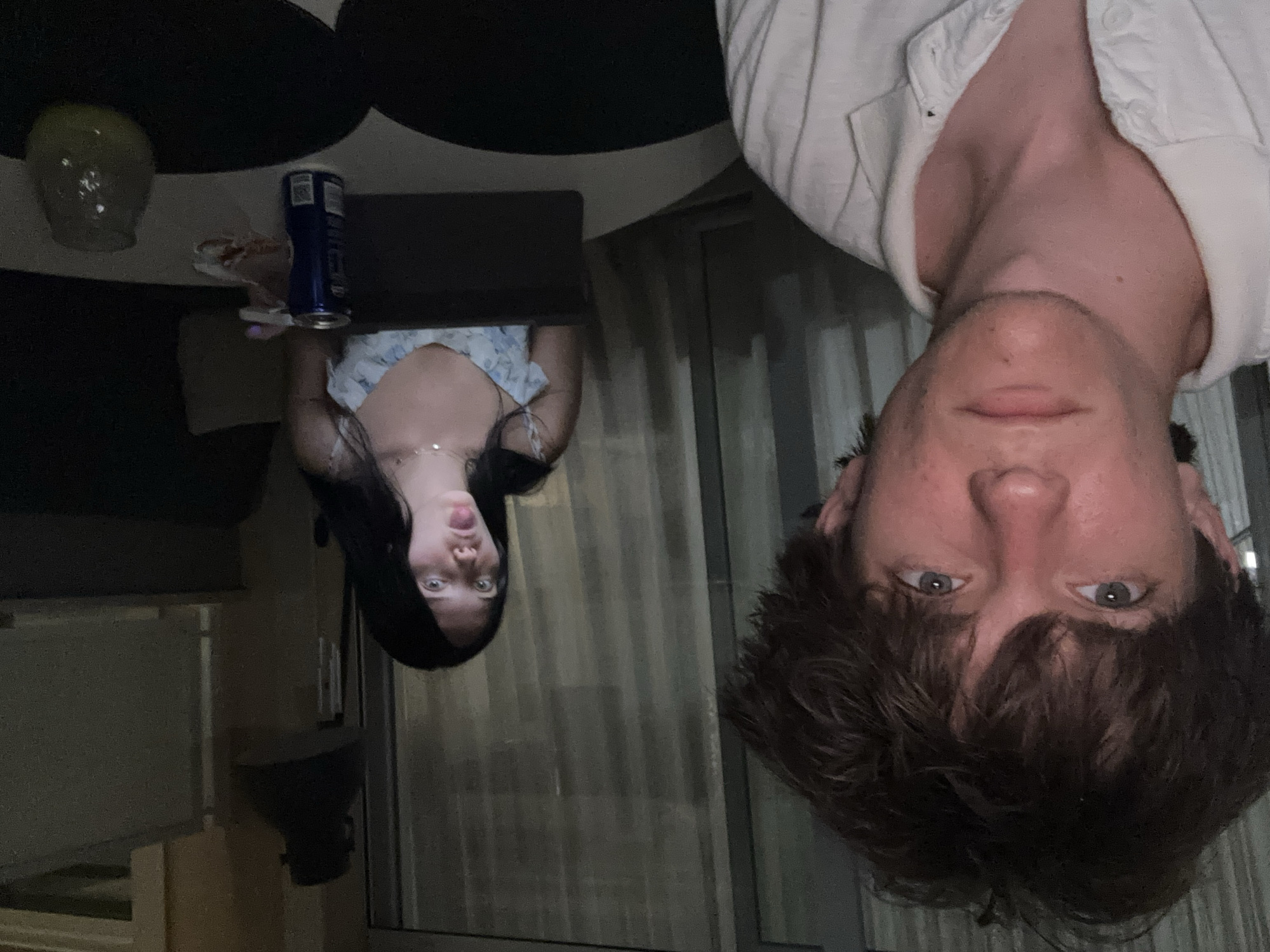 The image is a selfie taken by a young man with messy brown hair and light-colored eyes, wearing a white polo shirt. He is looking directly at the camera with a neutral expression. In the background, a young woman with long dark hair is visible, sitting at a table and sticking her tongue out at the camera. She is wearing a light-colored floral dress. On the table between them are a blue can and a glass with a yellow liquid. The scene appears to be indoors, with curtains visible in the background, suggesting a residential or hotel setting. The lighting is dim, with artificial light sources illuminating the subjects. This scene was likely captured in a residential balcony or patio area, potentially in Cupra Marittima, Italy, given the context provided.