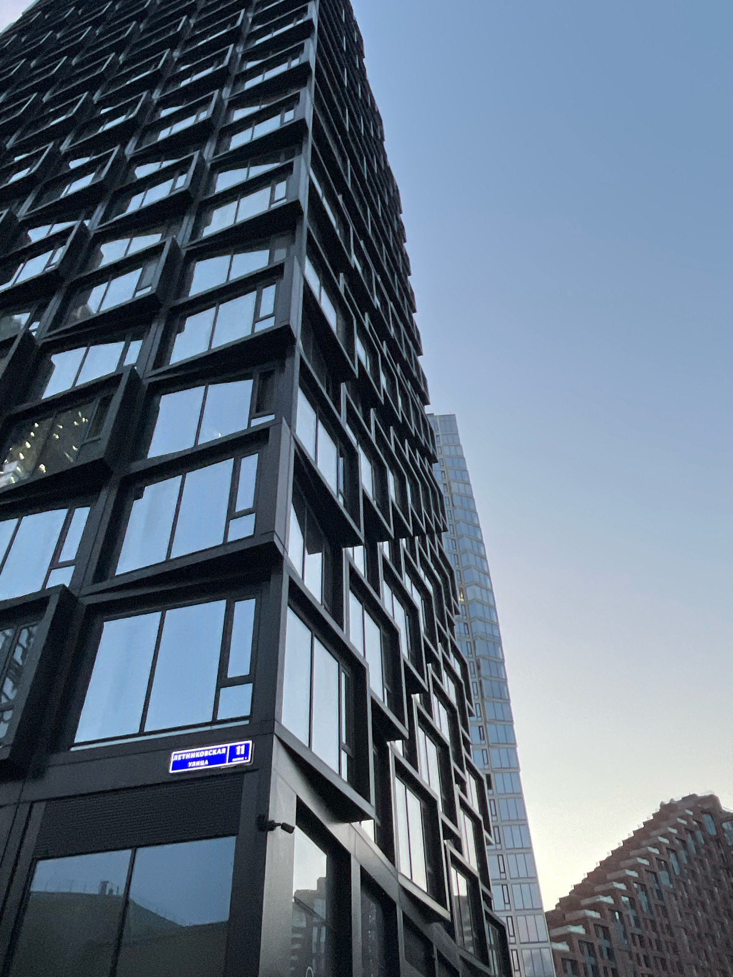 The image captures a low-angle view of modern, dark-colored high-rise buildings in Moscow, Russia, under a clear, pale blue sky. The primary focus is on a tall, black building with a distinctive facade featuring numerous recessed windows, creating a geometric, stepped appearance. To the right, another sleek, glass-covered skyscraper is visible, also extending upwards. In the far right, a building with a reddish-brown, textured facade and a sloped roofline is partially shown. A blue street sign with white text, "ЛЕТНИКОВСКАЯ 11", is affixed to the corner of the black building. No people are visible in the frame. The lighting suggests it is either early morning or late afternoon, with soft light illuminating the scene.