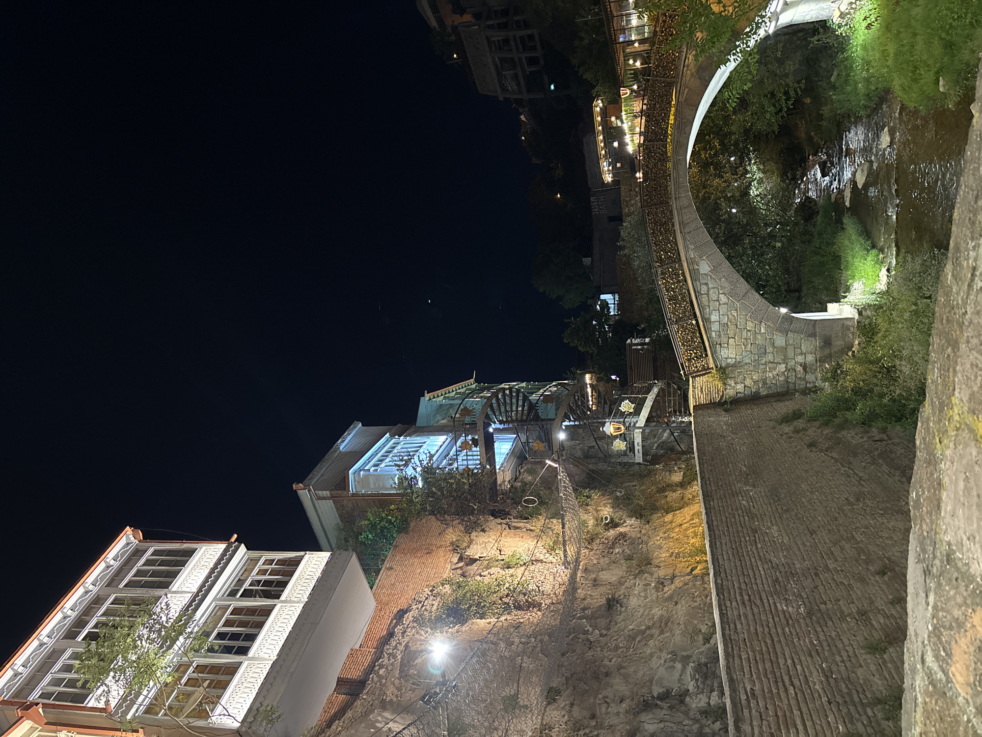 The image depicts a picturesque nighttime scene in a city, likely an urban area with historical architecture and a natural water feature. In the foreground, a rough stone embankment or walkway is visible. A stone arch bridge, adorned with numerous heart-shaped locks, spans over a shallow, rocky stream. The bridge is illuminated by warm lights along its railing, and lush green foliage and trees grow on its banks and underneath it.
On the left side of the image, a steep, rocky cliff face is illuminated by artificial lights, revealing a textured surface with patches of vegetation. Several structures are built into or on top of this cliff. The most prominent is a multi-story building with intricate white balconies featuring elaborate carvings and numerous windows. Below this, a section of brick wall and a net-like structure suspended on cables are visible against the cliff. Further up the cliff, another building with bright blue-lit windows and a spiral staircase attached to its exterior is present.
The background shows more buildings and structures on elevated terrain, also illuminated by lights, suggesting a densely populated area. The sky is dark, indicating it is nighttime. The overall impression is one of a charming and romantic urban setting, where nature and architecture are integrated.