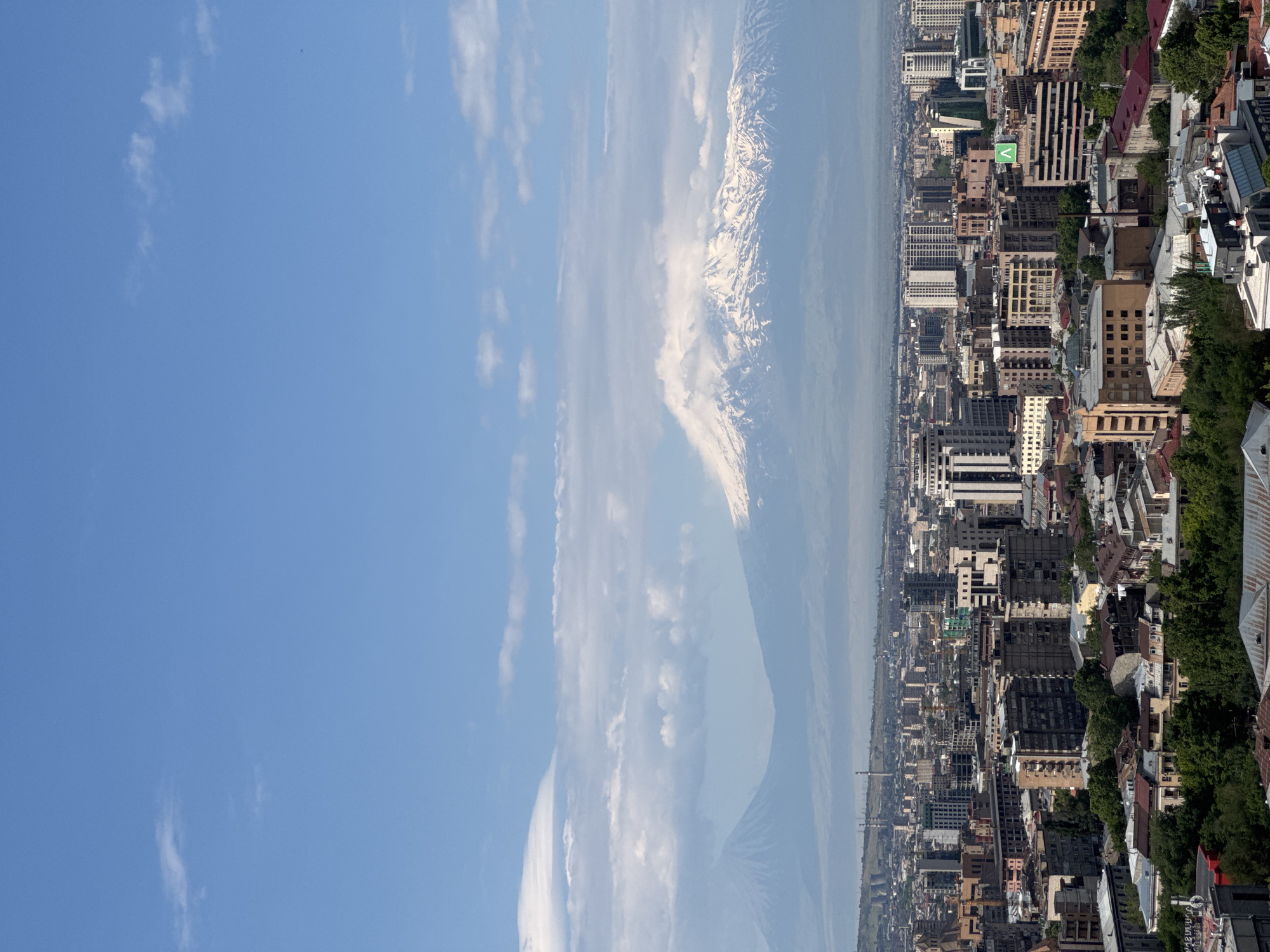 This panoramic image captures a sprawling city under a clear blue sky, with a majestic snow-capped mountain range visible in the distance. The perspective appears to be from an elevated viewpoint, looking out over a vast urban landscape.
The city, likely Yerevan, occupies the lower and mid-ground, showcasing a dense cluster of buildings. These structures exhibit a variety of architectural styles and heights, with many appearing to be multi-story residential or commercial buildings constructed from brick, stone, or concrete. Patches of green foliage, indicating trees or parks, are visible throughout the urban fabric, particularly towards the right.
Dominating the mid-ground and background on the left is a grand mountain, its upper reaches distinctly covered in bright white snow. Its iconic, imposing silhouette and snow-capped peaks strongly suggest Mount Ararat. Hazy atmospheric conditions lend a soft, ethereal quality to the lower slopes of the mountain, blending it with the horizon.
The sky above is a bright, clear blue, dotted with scattered, wispy white clouds that extend across the mountain and the upper left portion of the frame. The scene is illuminated by bright daylight, indicating good visibility.
Within the city, a notable detail is a green sign with a white 'V' or chevron-like logo visible on one of the buildings. No other text is clearly discernible. The overall impression is one of a vibrant city juxtaposed against a powerful and iconic natural landmark.