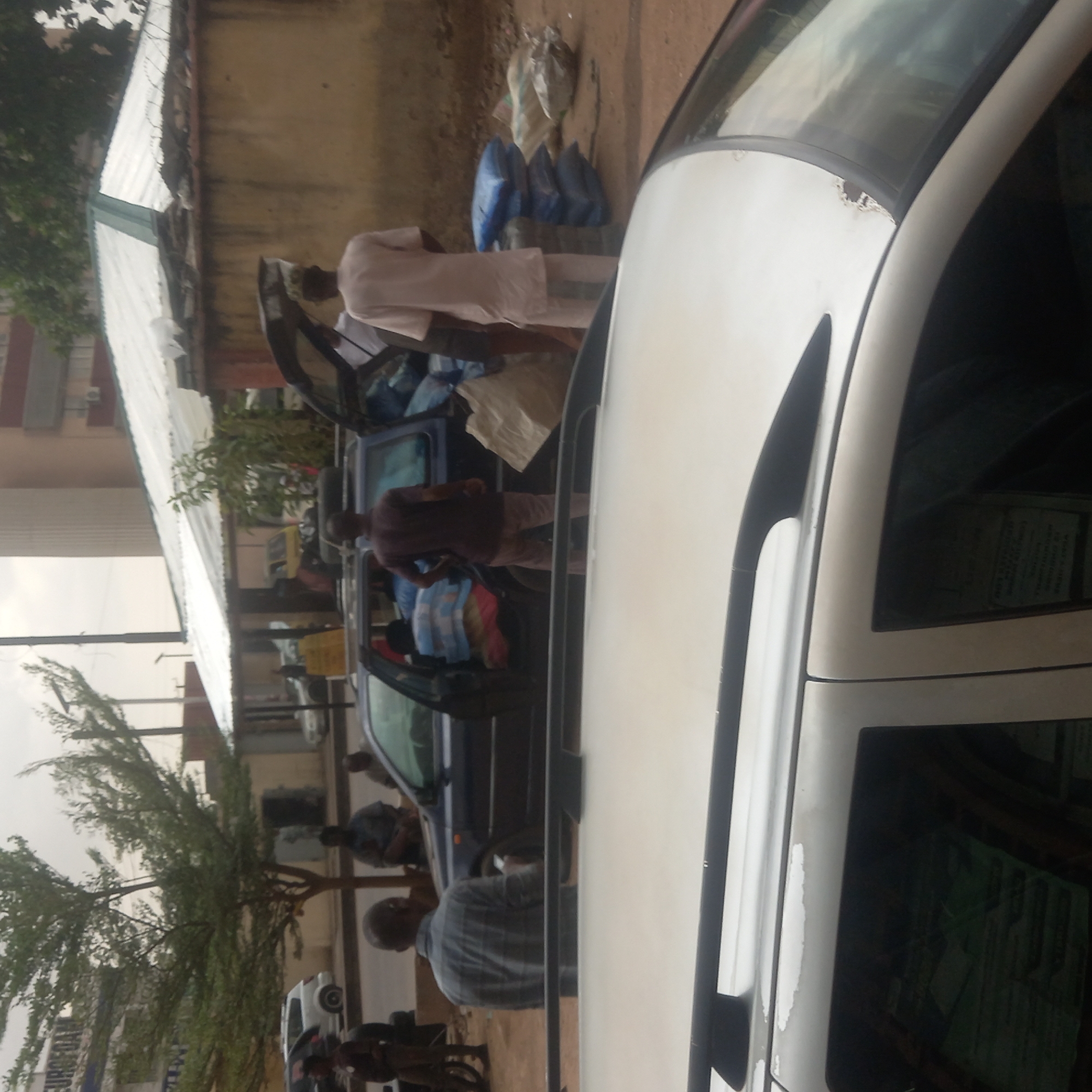 This media shows an outdoor daytime scene in what appears to be a street or parking area, consistent with the Dadi Riba, Nigeria context. The image is oriented vertically but depicts a horizontal setting.
The main activity involves several men interacting with at least two cars, primarily appearing to load or unload items. In the foreground on the right, a white car is partially visible. Notable details on this car include peeling paint on its roofline near the windshield. Reflected in its tinted windows is some unidentifiable, blurry text.
Towards the center-left, a dark blue or black car has its trunk open. A man wearing a light-colored, long-sleeved garment is bent over the open trunk, seemingly placing or retrieving items. Near him, several bags, some blue and others white or clear plastic, are visible on the ground. Another man in a dark reddish-brown shirt stands near the side of this dark car, and a third man in a plaid shirt is seen further back near an open door of the same car. Other people are visible in the background, some near other vehicles or walking.
The background features light-colored buildings, one with a corrugated metal roof. Lush green trees with thin branches and foliage are also prominent, suggesting an outdoor environment. The lighting suggests it is daytime, possibly overcast, with no harsh shadows. No clear emotions are discernible from the individuals, who appear focused on their tasks.
