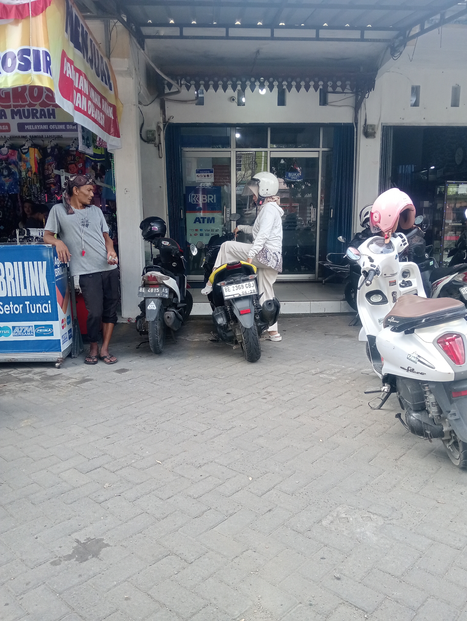 A street scene in Bandar Lampung, Indonesia, shows a man standing by a blue kiosk with the text "BRILINK Setor Tunai" on its side. He wears a gray t-shirt, dark pants, and flip-flops, with a bandana around his head. In front of him are two motorcycles. A woman in a white hoodie and pants, wearing a white helmet, sits on one of the motorcycles. Another motorcycle is parked nearby, and a woman in a pink helmet is also visible. The backdrop is a building with open storefronts, featuring signs for "ATM" and "BRI". Clothes are displayed for sale on racks to the left of the building. The ground is paved with interlocking bricks, and there are some puddles and debris present.