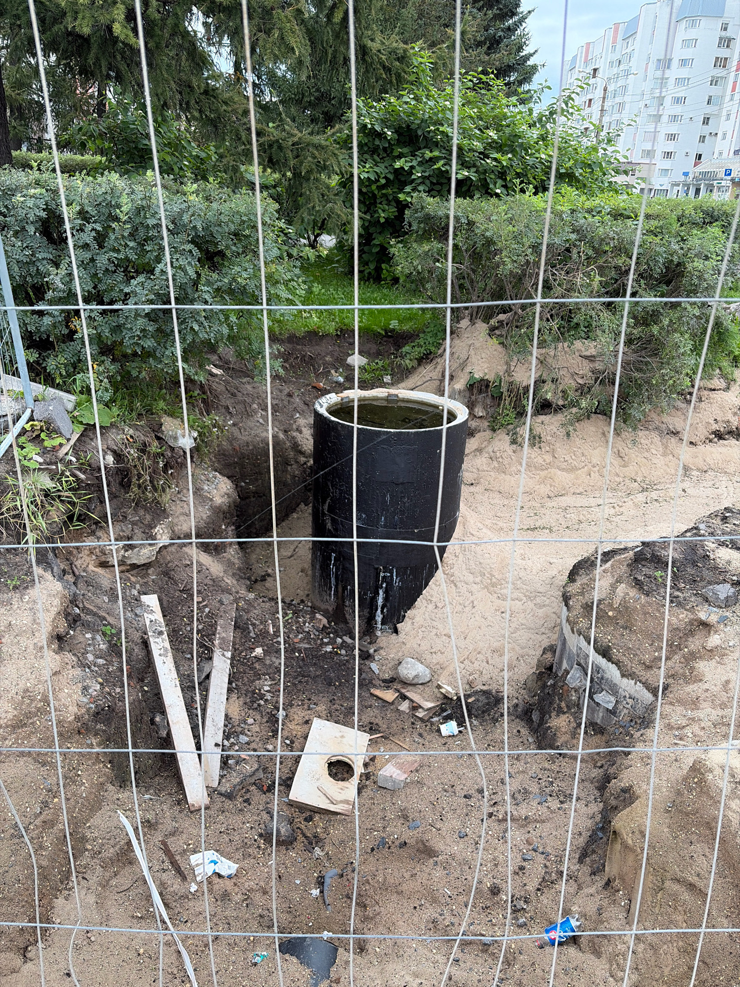 The image depicts an outdoor construction site in Arkhangelsk, Russia, as indicated by the context of the city and the presence of a modern apartment building in the background. The foreground is dominated by a wire mesh fence, which obstructs the view of the scene.
Inside the fenced area, there is a circular, black concrete pipe with a white rim, partially submerged in water. The pipe is situated in a dug-out area filled with sandy soil and some loose rocks. The surrounding ground is uneven, showing signs of excavation. Several pieces of construction debris, including wooden planks, concrete fragments, and a small square wooden board with a circular opening, are scattered around the site. There are also some plants and green foliage growing in the background, suggesting a park or green space adjacent to the construction area. The sky is overcast. No people are visible in the image. The scene appears to be a municipal construction project, possibly related to underground utilities or infrastructure.