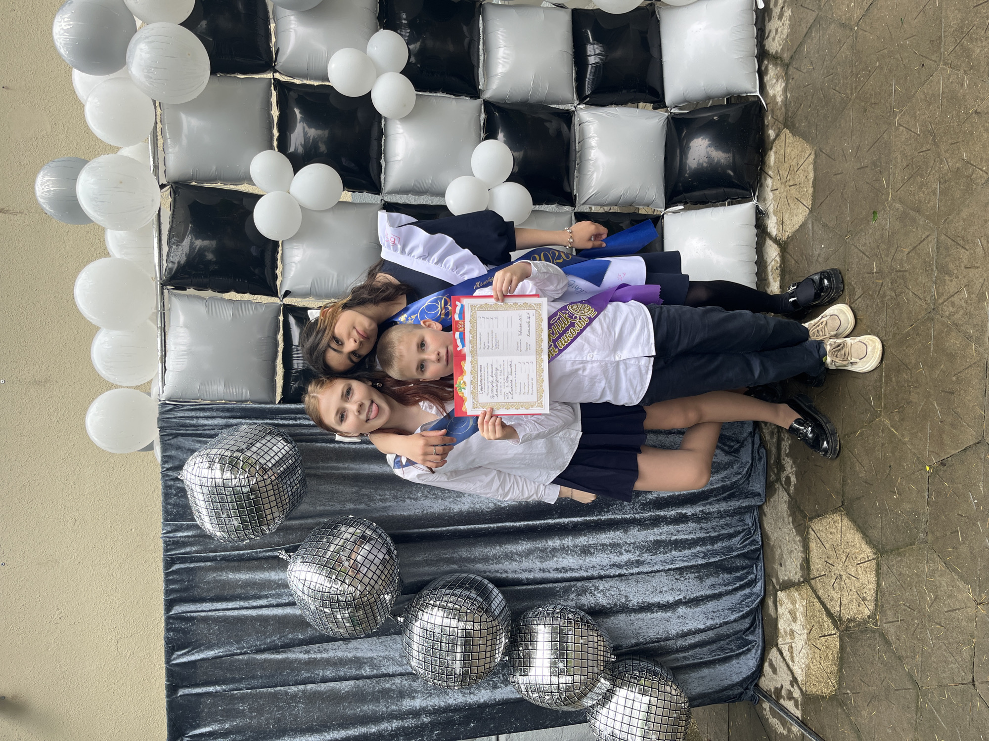 Three individuals, possibly siblings, are posed on a gray velvet cloth, situated on an outdoor paved surface. The children appear to be celebrating a graduation or achievement, as one holds a certificate. The backdrop consists of alternating black and white square cushions, adorned with clusters of white balloons. Five disco balls are placed on the velvet cloth in front. The pavement surface is slightly wet. The photo was taken in Nizhnyaya Shilovka, Russia.