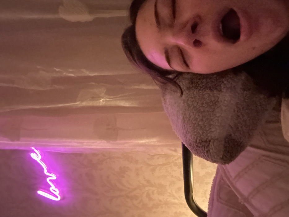 A person with dark hair and closed eyes is positioned at the top of the frame, mouth open in a yawn. They rest against a plush, textured surface. Below, a patterned wallpaper is visible, illuminated by a pink "love" neon sign on the left. A sheer curtain hangs in the background. A dark metal rod is also present, suggesting a furniture or bed frame. The overall lighting creates a warm, relaxed atmosphere.