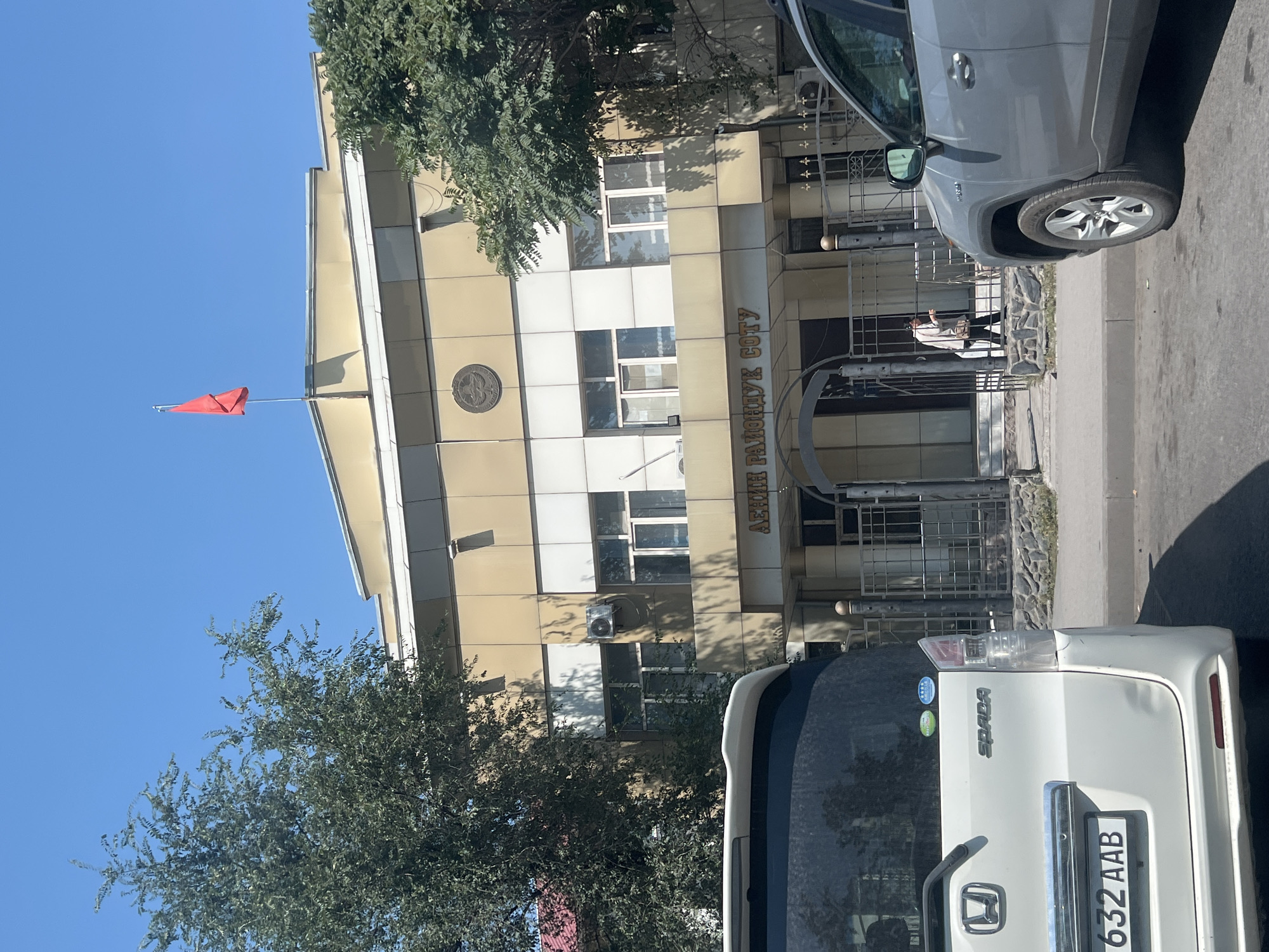 In Bishkek, Kyrgyzstan, a photograph captures the front of a building, likely a government or public institution, with a red flag atop its roof. The building has a light-colored facade with metallic accents and rows of windows. Below the main entrance, text is visible in Cyrillic, reading "ЛЕНИН РАЙОНДУК СОТУ," which translates to "Lenin District Court." A woman in dark clothing is seen walking towards the building's entrance. In the foreground, the rear of a white Honda Spada minivan and the side of a silver Toyota RAV4 are visible, parked on a street adjacent to the building. The scene is set under a clear blue sky, with trees partially obscuring the building's sides.