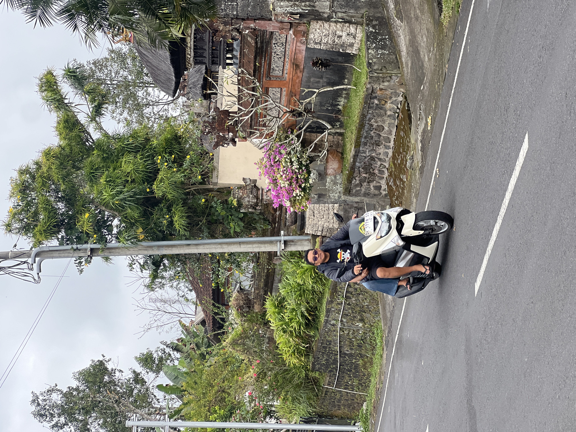 Two individuals are riding a white scooter on a paved road in Rendang, Indonesia. The person in the front is wearing sunglasses, a black t-shirt with a graphic print, and black shorts. The person in the back is wearing a blue garment and dark flip-flops. The scooter has a license plate that reads "DK 5778 SD". The background features lush greenery, a stone wall, and traditional Balinese architecture with thatched roofs. The sky is overcast.