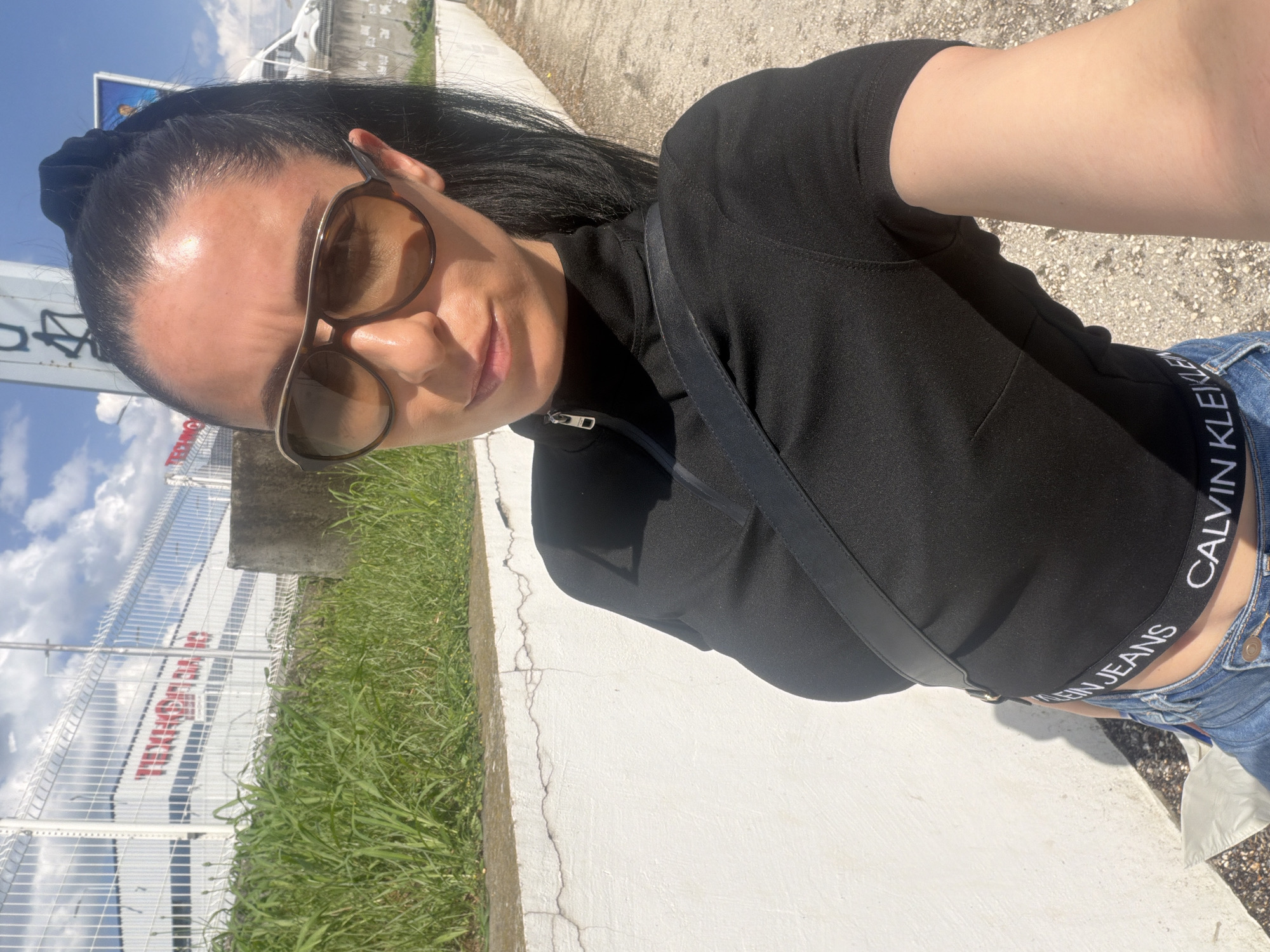 A medium-shot, canted photograph of a woman outdoors, likely taken by herself via an outstretched arm. She has dark hair pulled back, wears sunglasses, a black top, a shoulder bag strap, and blue jeans with a visible Calvin Klein waistband. The background includes a concrete barrier, grass, a metal fence, and a building with red and white signage, indicating a business. The sky is visible in the upper-left, showing partly cloudy conditions. The location can be identified as Pleven, Bulgaria, due to the signage on the building behind the metal fence.