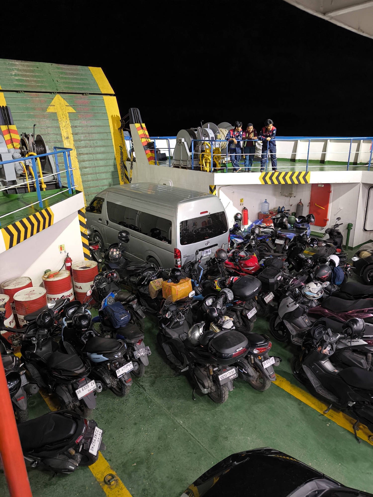 The image depicts the interior of a ferry or large boat, possibly a vehicle ferry. The location of the city is unknown. There are several motorcycles parked tightly together on the green-colored deck. Also parked on the deck is a silver van. The deck has yellow stripes marking parking areas and pathways. There are large red drums with white lettering in the foreground.
On an upper deck in the background, three people stand looking out over the scene. The sky is dark, suggesting it is either night or the interior of the ferry is poorly lit. There are various pieces of equipment and safety devices visible, such as fire extinguishers.