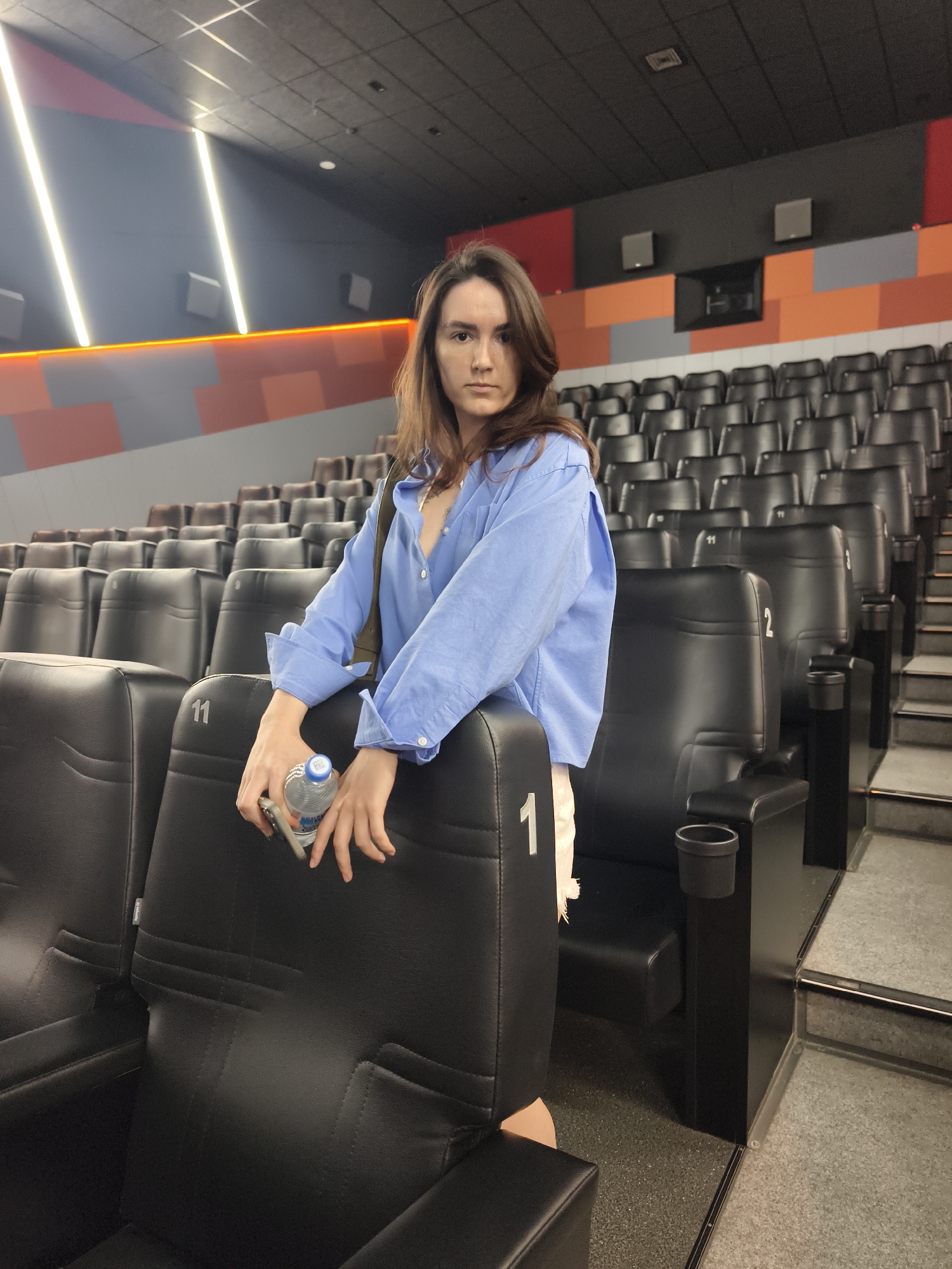 A young woman with long brown hair is leaning on a row of empty black theater seats in a movie theater. She is wearing a light blue button-down shirt and white pants. She is holding a water bottle and a mobile phone in her hands. The seats are arranged in tiered rows. The walls of the theater are decorated with a pattern of orange, gray, and red rectangular shapes, and illuminated by vertical strip lights. The floor is carpeted with a gray speckled material. The scene is indoors. The image was taken in Tyumen, Russia.