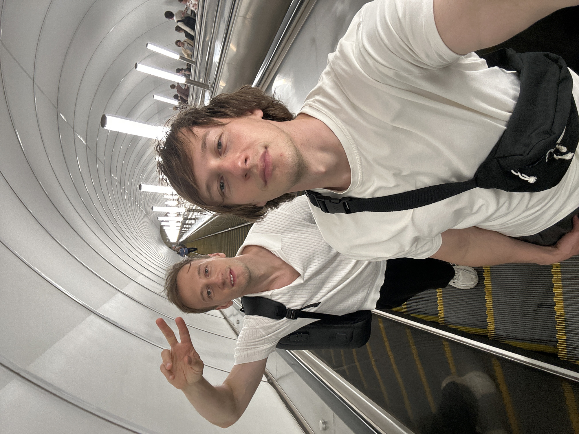 The image shows two young men on an escalator, likely in a subway station in Saint Petersburg, Russia. The man in the foreground is closer to the camera and is wearing a white t-shirt and black pants, with a black fanny pack around his waist. He is looking at the camera and holding up two fingers in a peace sign. The other man is further down the escalator and is also wearing a white t-shirt and black pants. He is partially obscured by the first man. The escalator has yellow safety lines on the steps. The background shows the curved, tunnel-like architecture of the escalator shaft, with rows of fluorescent lights. The time of day is not discernible, and the weather is irrelevant as the scene is indoors. The overall mood seems casual and friendly. There is no visible text in the image.