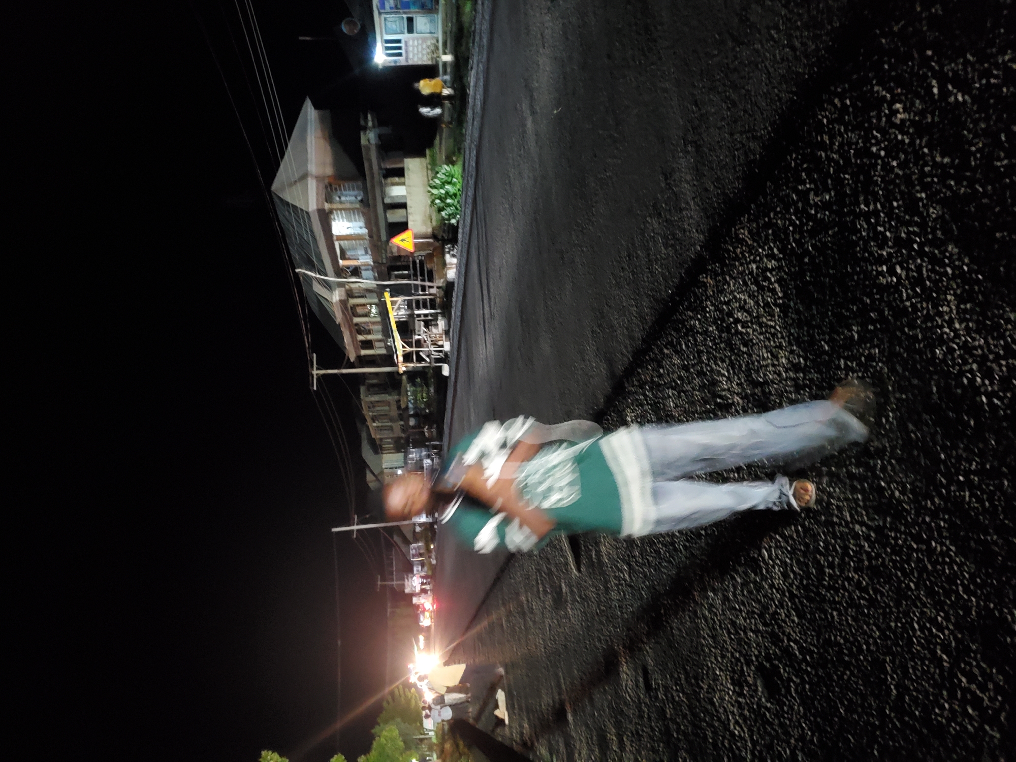 This is a night-time photograph taken outdoors in Jalingo, Nigeria, likely along a paved road. In the foreground, a person is walking towards the camera, appearing slightly blurred due to motion. They are wearing a green and white striped shirt and light-colored pants. The person is carrying a white object, possibly a bundle of cloth, in their arms. To the left of the foreground figure, several other individuals are standing on the side of the road, also appearing somewhat blurred. They are near some trees. In the background, there are buildings, some of which are illuminated. Streetlights and vehicle headlights are visible in the distance, indicating traffic. The road surface appears to be asphalt, with a gravel shoulder on the left. Power lines are visible stretching across the frame. The overall scene depicts a street at night in a Nigerian city.