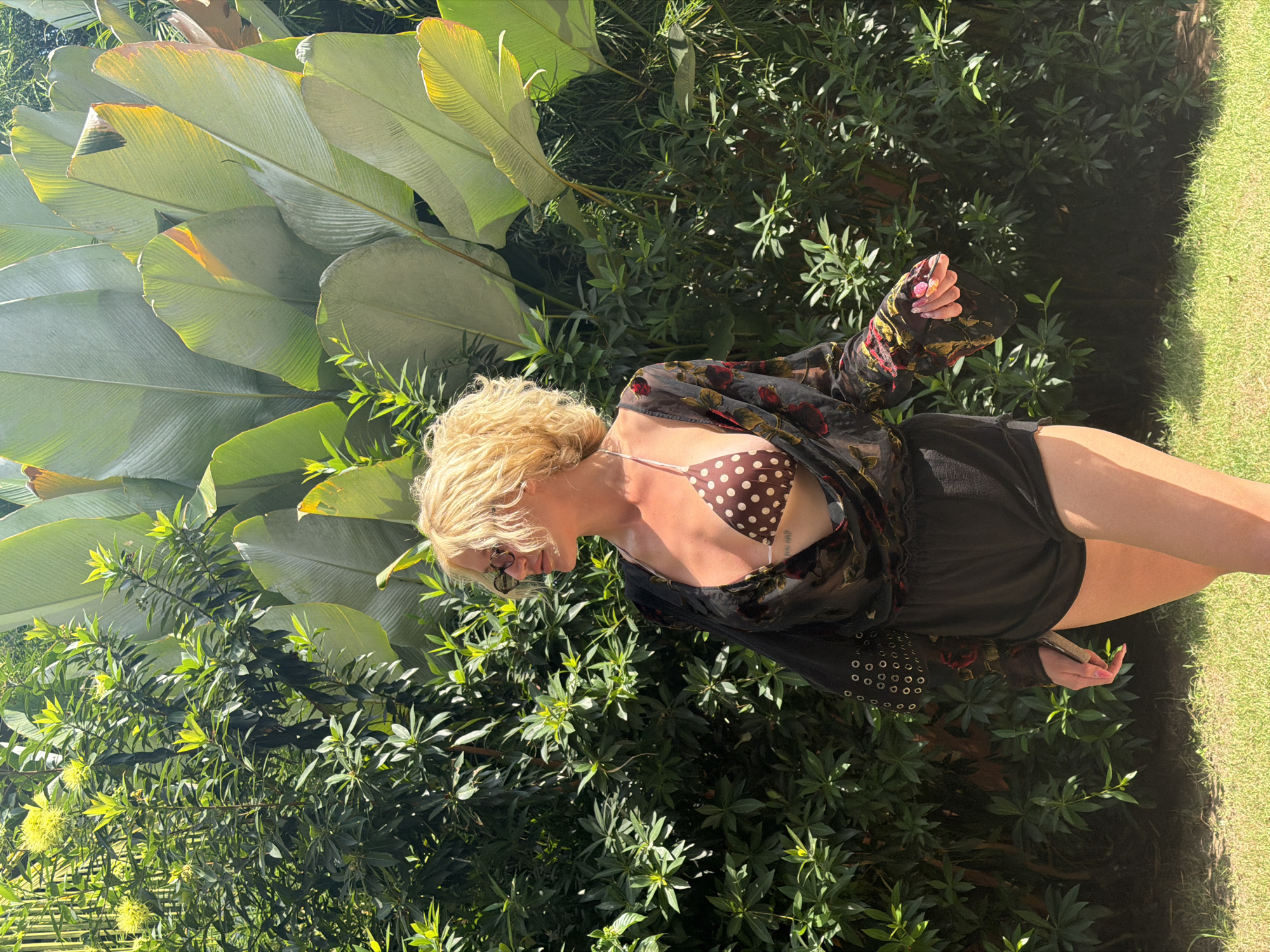 A blonde woman with curly hair, wearing sunglasses, a brown polka dot bikini top, a sheer black floral printed cover-up, and black shorts, stands in front of lush green foliage and large tropical leaves. She is looking to her right. The scene appears to be outdoors in a tropical setting, potentially in North Kuta, Indonesia, given the dense vegetation and large leaves suggestive of a humid climate. The ground is covered in green grass.