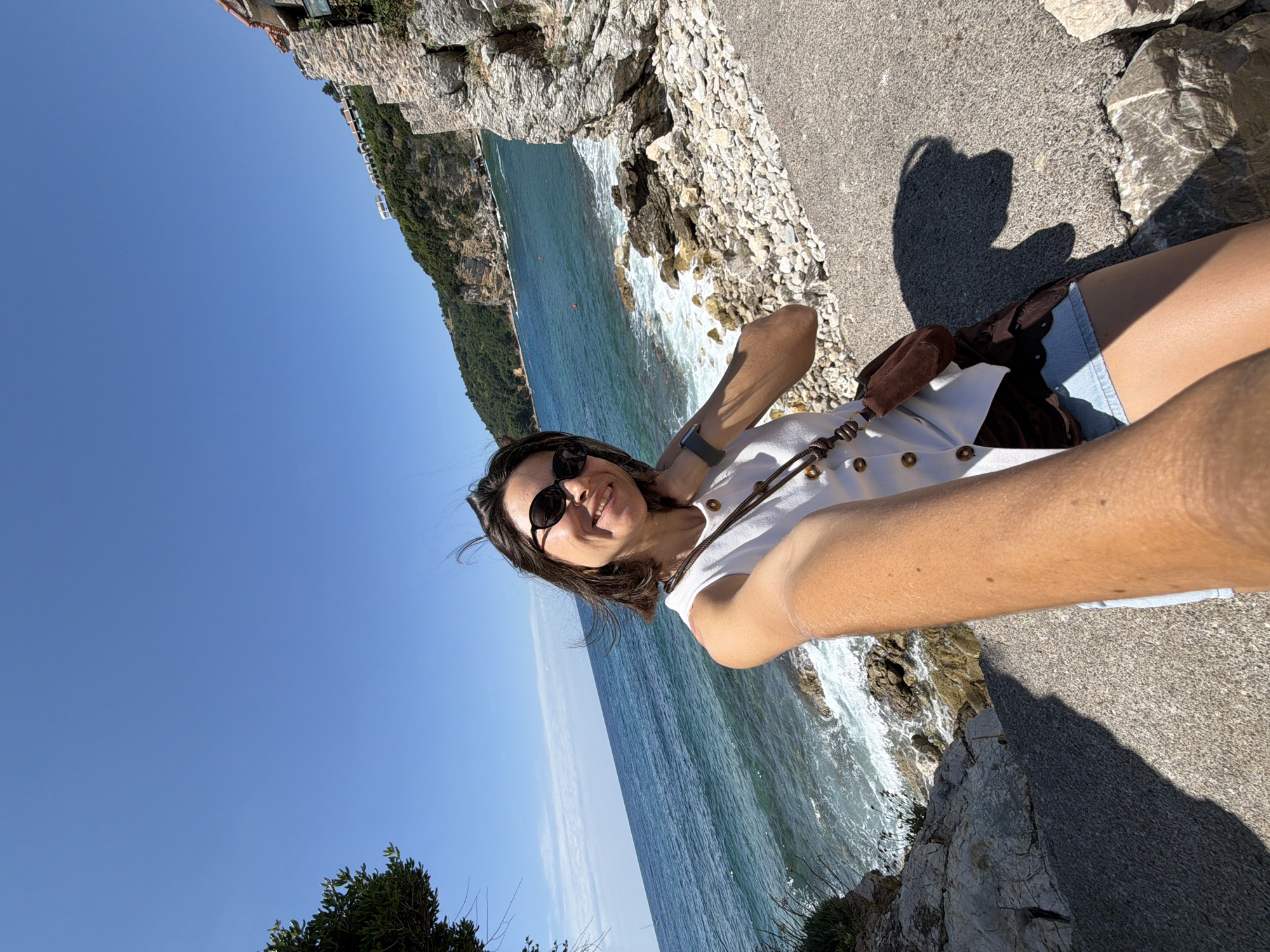 A woman is captured in a cheerful selfie, smiling broadly at the camera while standing outdoors by a sunny coastline. She wears dark sunglasses, a white sleeveless top adorned with gold-toned button-like embellishments, and light blue denim shorts. A small brown shoulder bag is visible on her lap, and a dark-strapped smartwatch is on her left wrist.
The background features a picturesque coastal landscape. To her left, the clear blue-green sea stretches out under a bright, cloudless sky, with white waves breaking against the rugged, light-colored rocky shore. A steep, lush green hillside, dotted with buildings, extends into the distance. She is positioned on a paved or gravel path that runs along the edge of the rocks. The strong sunlight creates prominent shadows, indicating a bright, clear day. The scene evokes a sunny holiday atmosphere in a Mediterranean setting, consistent with Budva, Montenegro. No discernible text is visible in the image.