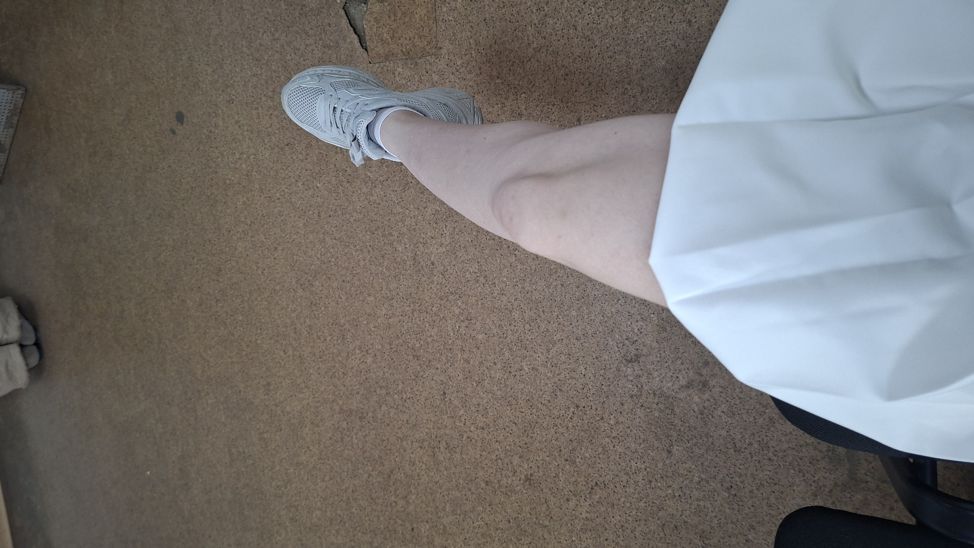 The image depicts a close-up view of a person's legs and feet, resting on a cork-like floor. The person is wearing light gray athletic shoes with white laces and socks. One leg is bent at the knee, with the foot extended. A large expanse of white fabric, possibly a skirt or sheet, covers the lower part of the person's body and a portion of the chair they are seated in, which is partially visible on the left side of the frame. The floor has a speckled brown texture. There is no discernible urban landscape or specific Chaikovsky location visible in this image.