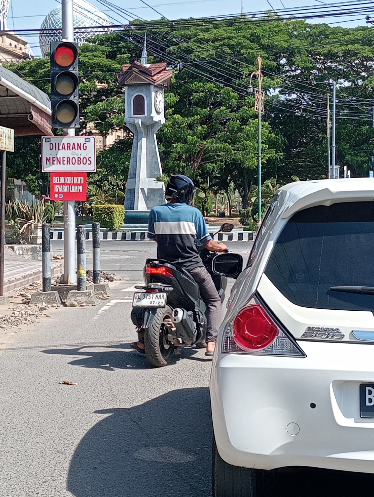 The image depicts a street scene in Lhokseumawe, Indonesia. A red traffic light is illuminated, and a sign with Indonesian text ("DILARANG MENEROBOS BELOK KIRI IKUTI ISYARAT LAMPU") is visible, which indicates it is prohibited to run the red light and to turn left following the signal lamp. A man on a motorcycle with the license plate "BL 3151 NAR" is positioned at the intersection, partially obscured by the rear of a white Honda Brio car. A clock tower stands in the background amidst green trees. The motorcycle's license plate also indicates the date "07-29".