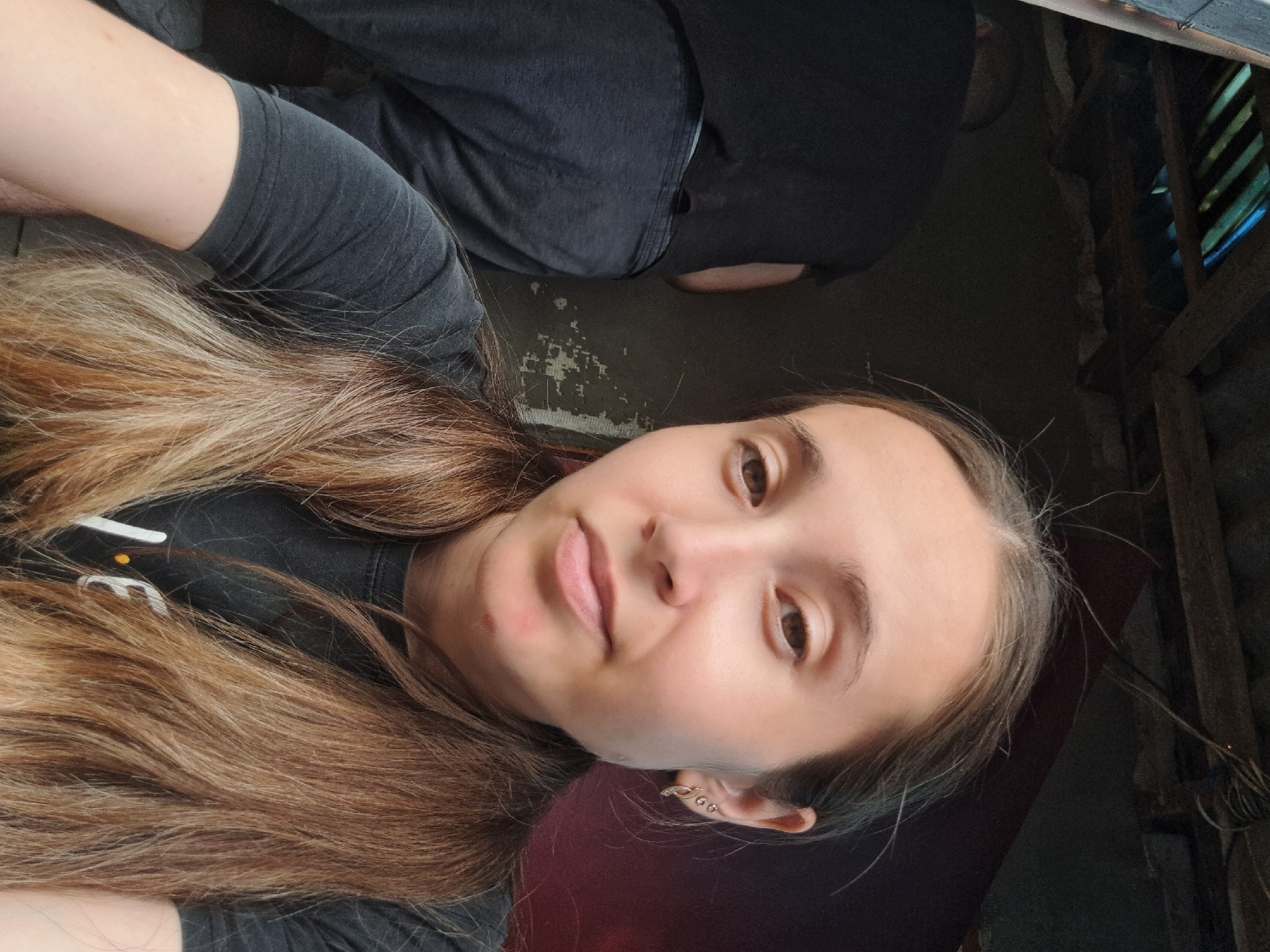 A selfie is taken by a young woman with light brown hair in two braids, wearing a dark t-shirt. She is in the foreground, with her face and upper body filling most of the frame. Behind her, to the left, a person wearing a black t-shirt and dark pants is visible from the back, facing away from the camera. The environment appears to be an indoor, unfinished space with a rough wooden ceiling and exposed beams. The walls are textured and show signs of wear, with peeling paint or plaster. The overall lighting is dim. The location is identified as the city of Vypasne, Ukraine.