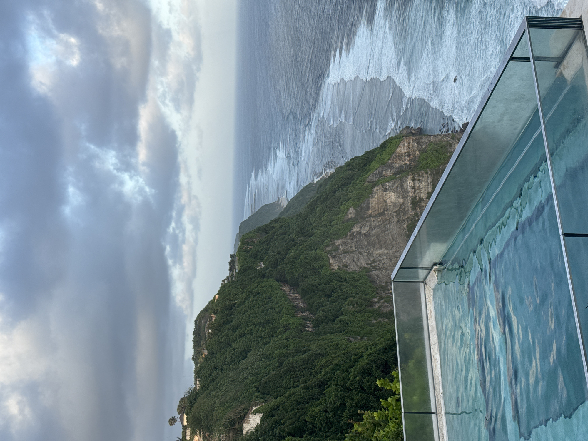 The image depicts a coastal landscape with a dramatic cliffside view. A glass-walled infinity pool is positioned on the edge of the cliff, creating the illusion of the pool water merging with the ocean. The cliff is covered in lush green vegetation and rocky outcrops. The ocean stretches out to the horizon with waves crashing along the shoreline. The sky is overcast with grey and white clouds. Location: Unknown, Unknown.