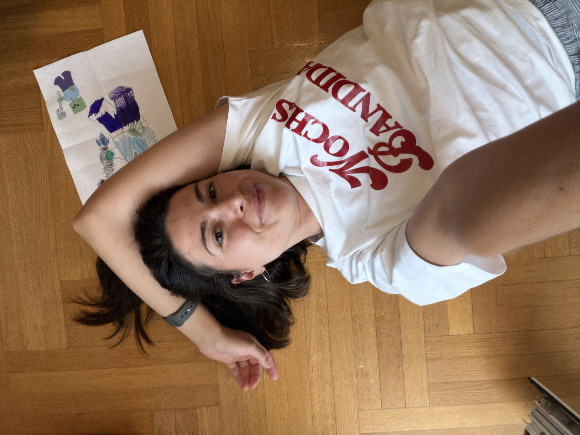 A woman is lying on a wooden floor. She is wearing a white t-shirt with red writing. Next to her is a child's drawing on white paper. The scene is indoors, and the light is natural. The city of Budva, Montenegro, is not discernible from the visual information provided.