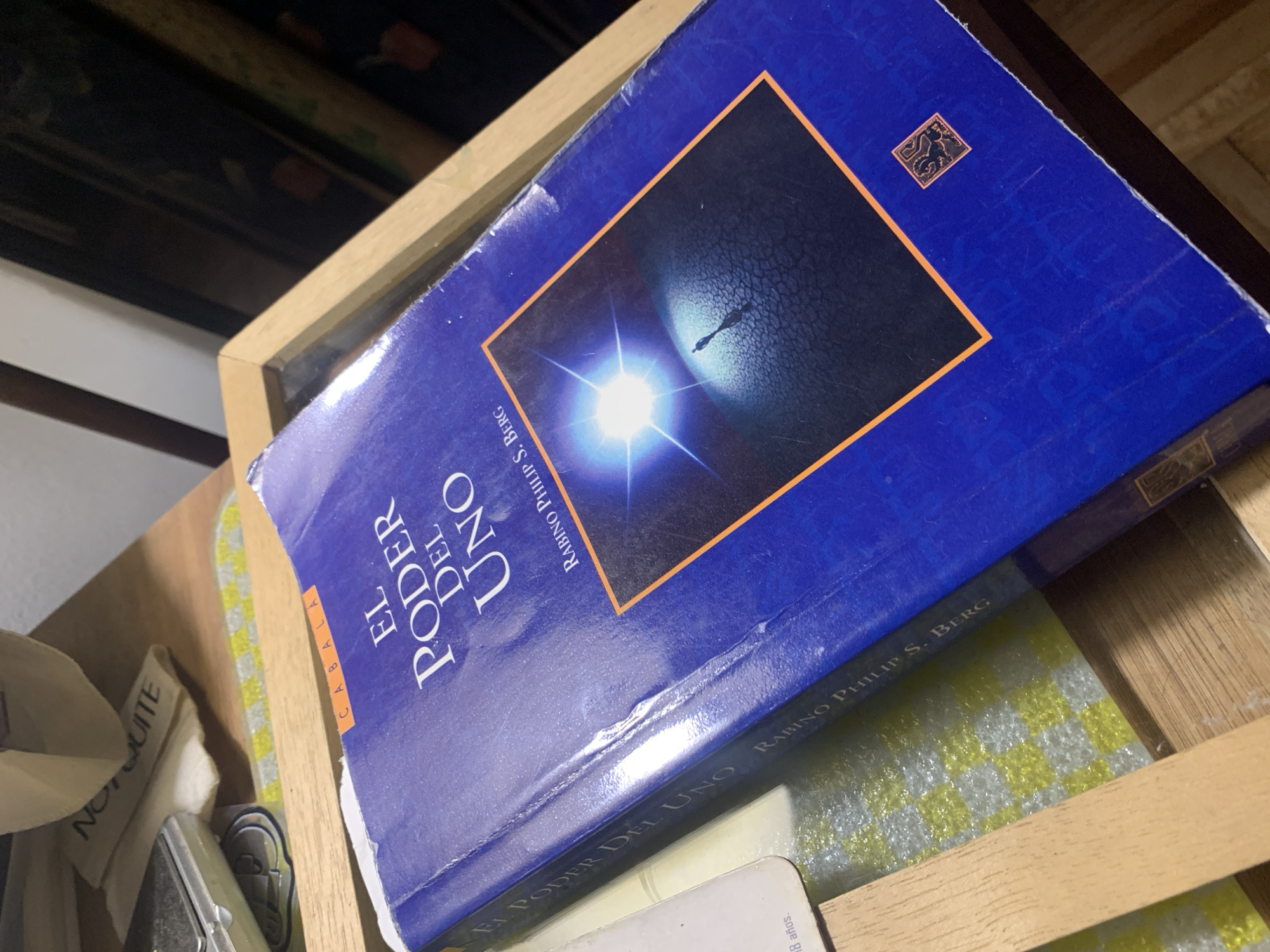A book titled "El Poder Del Uno" by Rabino Philip S. Berg is visible in the image. The book has a blue cover with a square image enclosed in an orange border. The square image shows a dark landscape with a bright light and figure. The book is in a wooden frame, likely on a shelf. Based on the text visible ("El Poder del Uno") it can be confirmed that the book is written in Spanish, indicating that the image was taken in a Spanish-speaking location. The title "EL PODER DEL UNO" hints that the book may have been written in Argentina or Buenos Aires, as this book is sold in this location. The image is taken at close range.