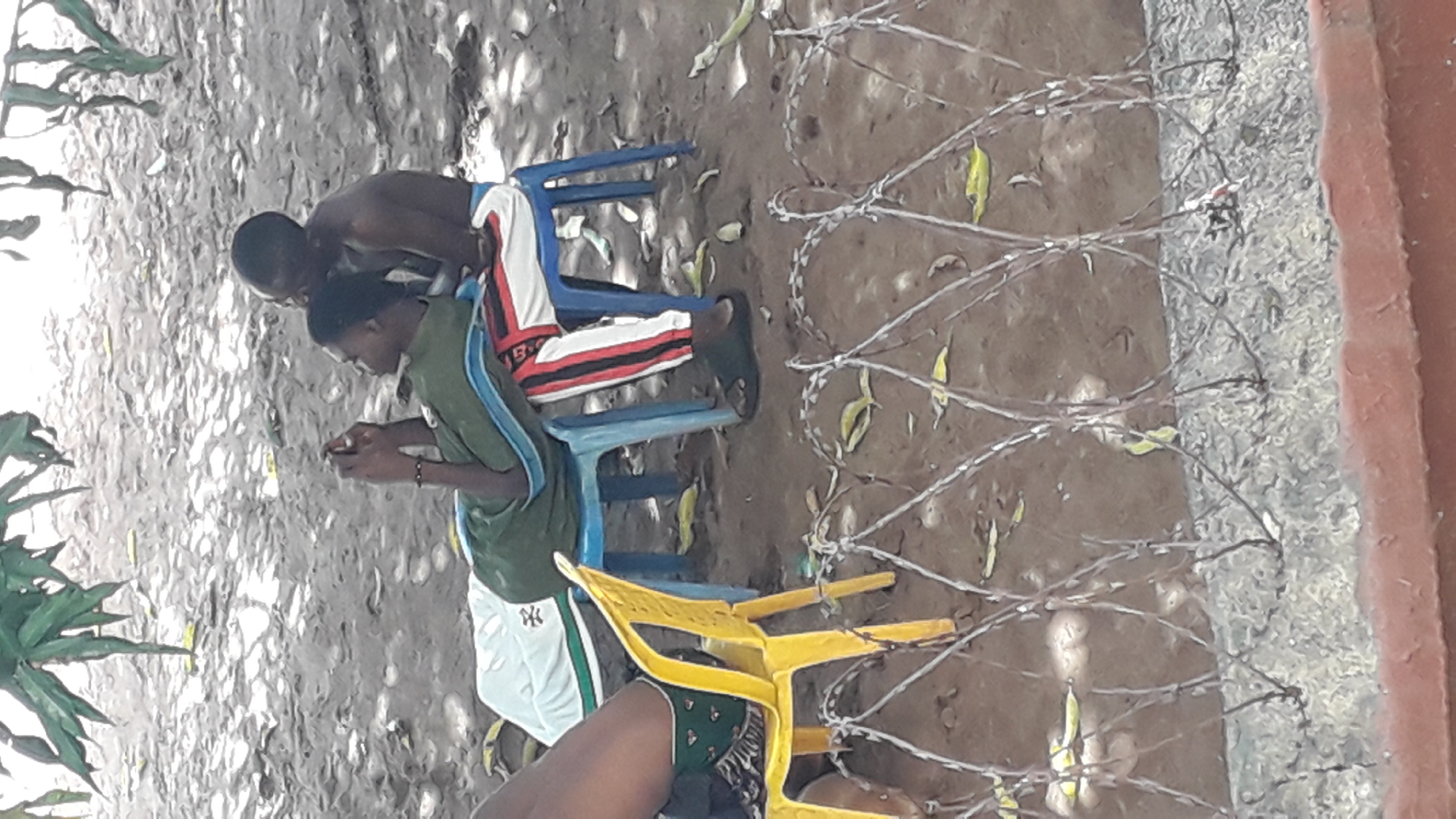 The image depicts two young men in an outdoor setting in Nkpor, Nigeria. One man is shirtless, wearing shorts, and sits on a blue plastic chair. The other man, wearing a green shirt and shorts, sits next to him, looking at a mobile phone in his hands. A yellow plastic chair is visible in the foreground. Barbed wire runs along the right edge of the frame. The ground is bare, with some scattered leaves. A portion of a building is visible on the right edge. Foliage is visible on the left side of the frame.