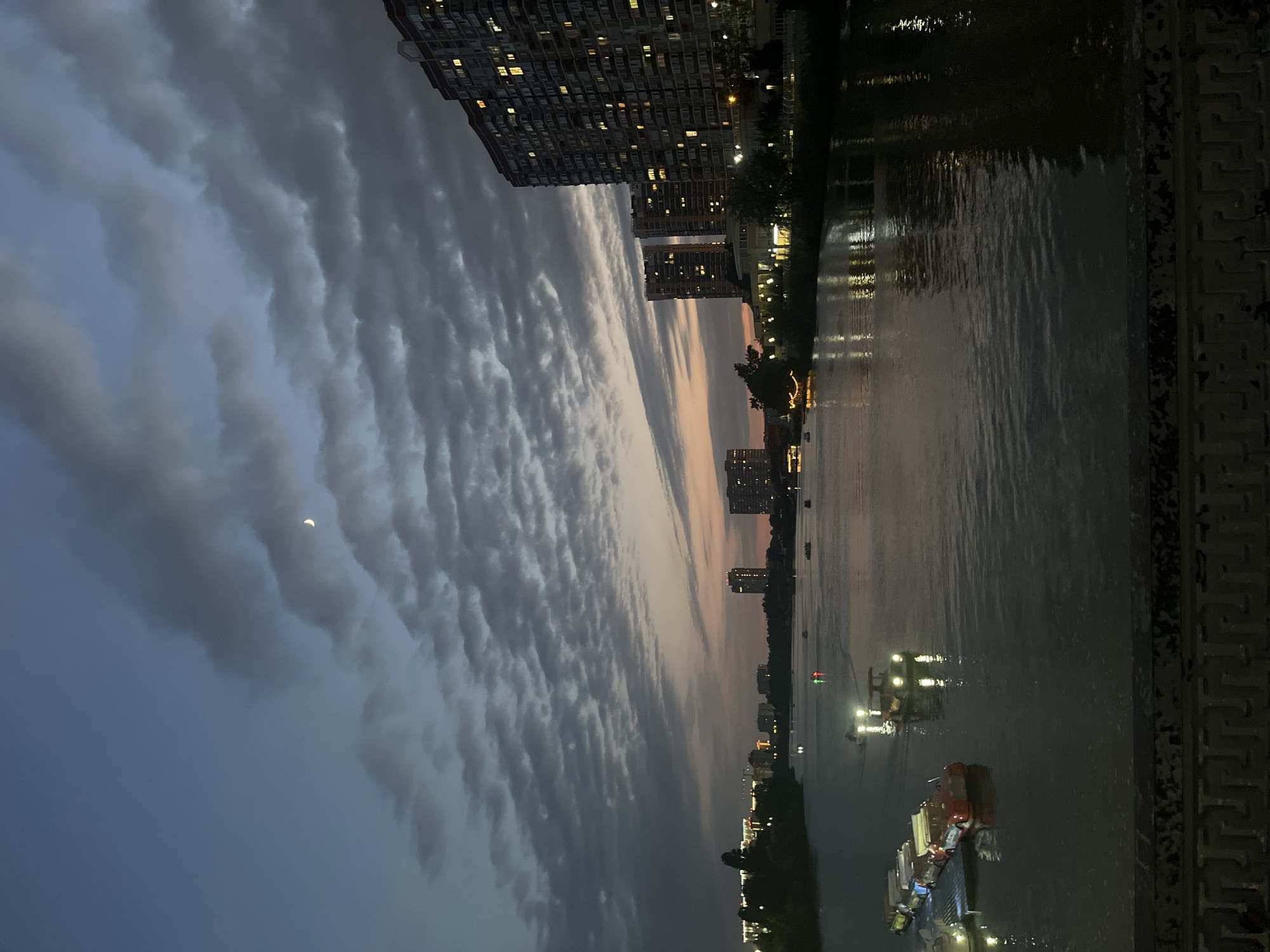 The image depicts a twilight scene over the Kuban River in Krasnodar, Russia. The sky is a gradient of deep blue and fading orange, with scattered, textured clouds. A sliver of the moon is visible in the upper left quadrant of the sky.
On the right side of the river, a row of tall, multi-story apartment buildings stands silhouetted against the sky. Their windows are illuminated with warm, yellow light, indicating occupancy. A shoreline with trees and lights is visible along the riverbank in front of the buildings.
The left side of the river shows a more distant cityscape with several buildings, some of which are illuminated.
The river itself is dark and reflective, with gentle ripples. Several small boats are scattered across the water. In the foreground on the left, a line of brightly colored pedal boats are docked, some with their lights on. Further out on the water, other boats, some with lights, are moving. The reflections of the lights from the boats and the shoreline are visible on the water's surface.
In the very bottom of the image, a dark, ornate railing or barrier is visible, suggesting the photograph was taken from a bridge or elevated walkway. The overall atmosphere is calm and serene, with the urban environment transitioning into the evening.