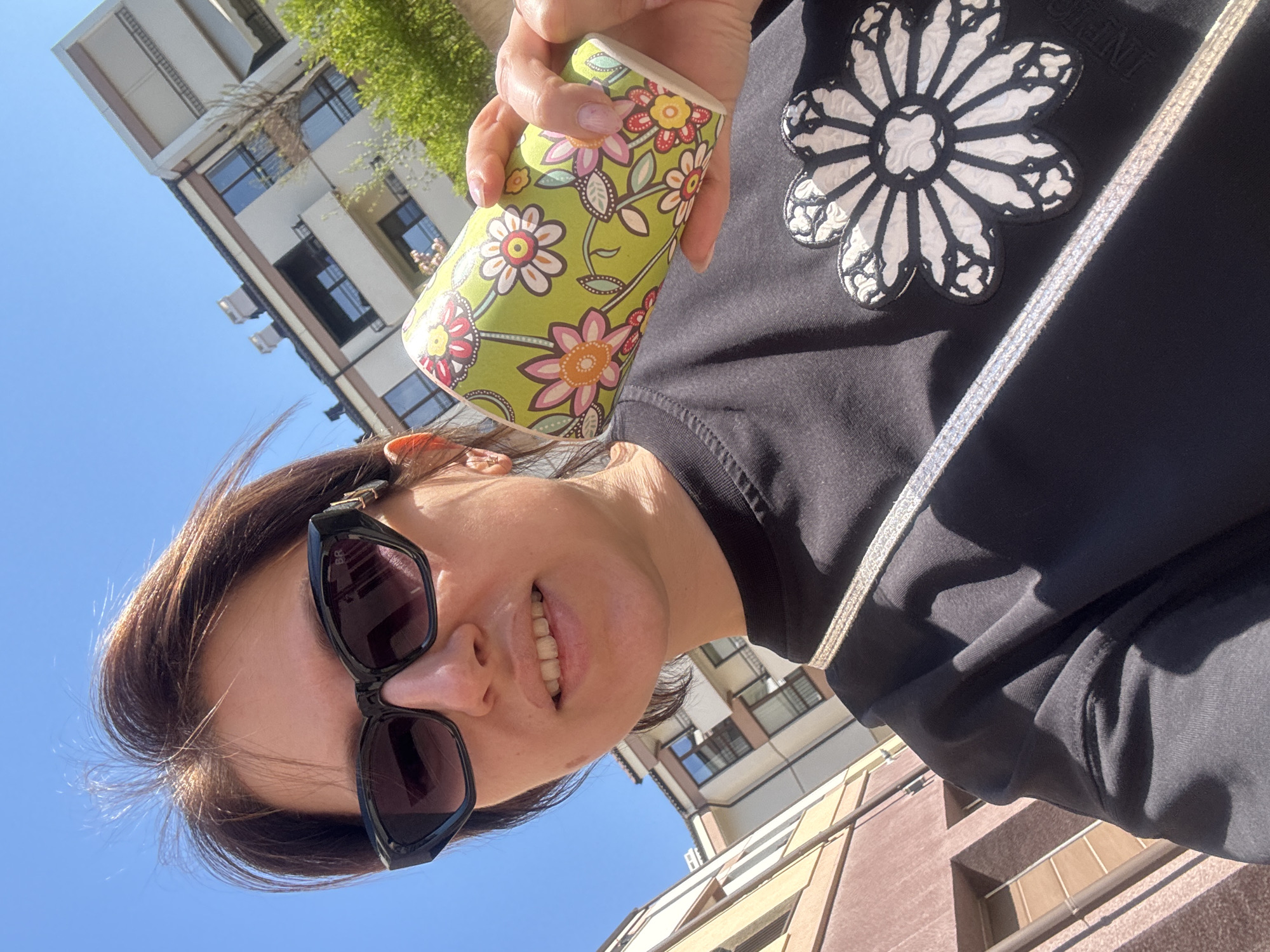 A person with dark hair and sunglasses holds a green and floral-patterned cup in their right hand, wearing a black long-sleeved shirt with a large flower design on the chest. The background features a clear blue sky and buildings with windows, suggesting an urban environment. Based on the architectural style and available information, the location could be identified as Sofia, Bulgaria. The person appears to be taking a selfie.