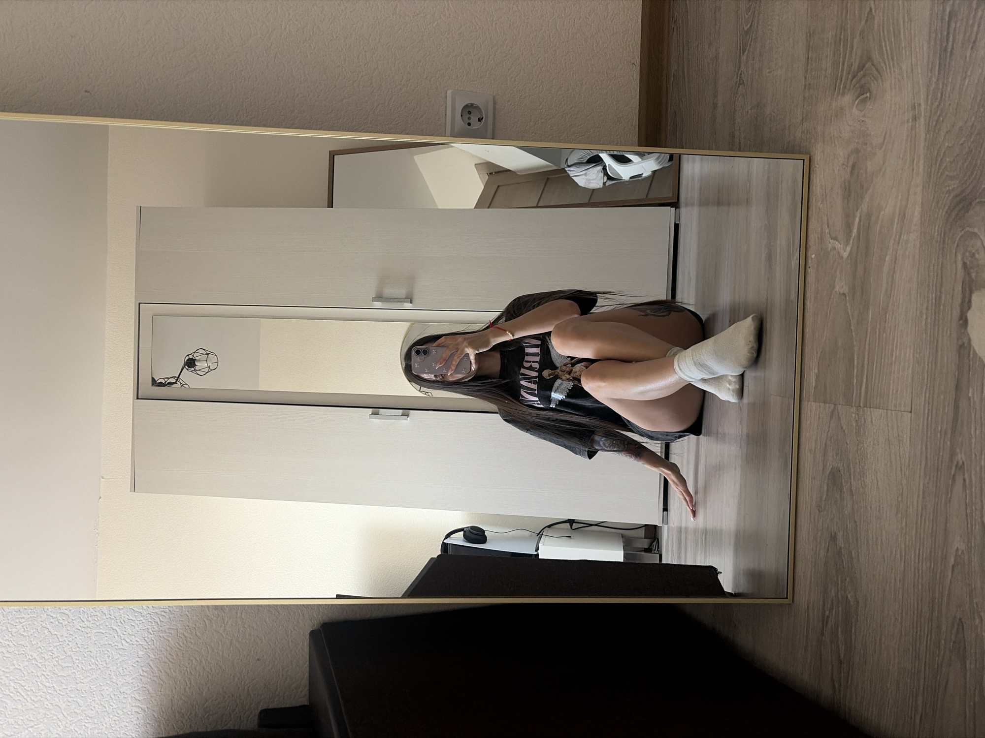 A young woman is reflected in a full-length mirror. She is sitting on the floor and taking a selfie with her phone. She is wearing a black t-shirt, dark shorts, and white socks. Her dark hair is long and straight. A black sofa is visible in the foreground. In the background, there is a white wardrobe, a wall outlet, and a wooden floor. The location is likely inside an apartment or house. The city, Saint Petersburg, Russia, cannot be determined from the image.